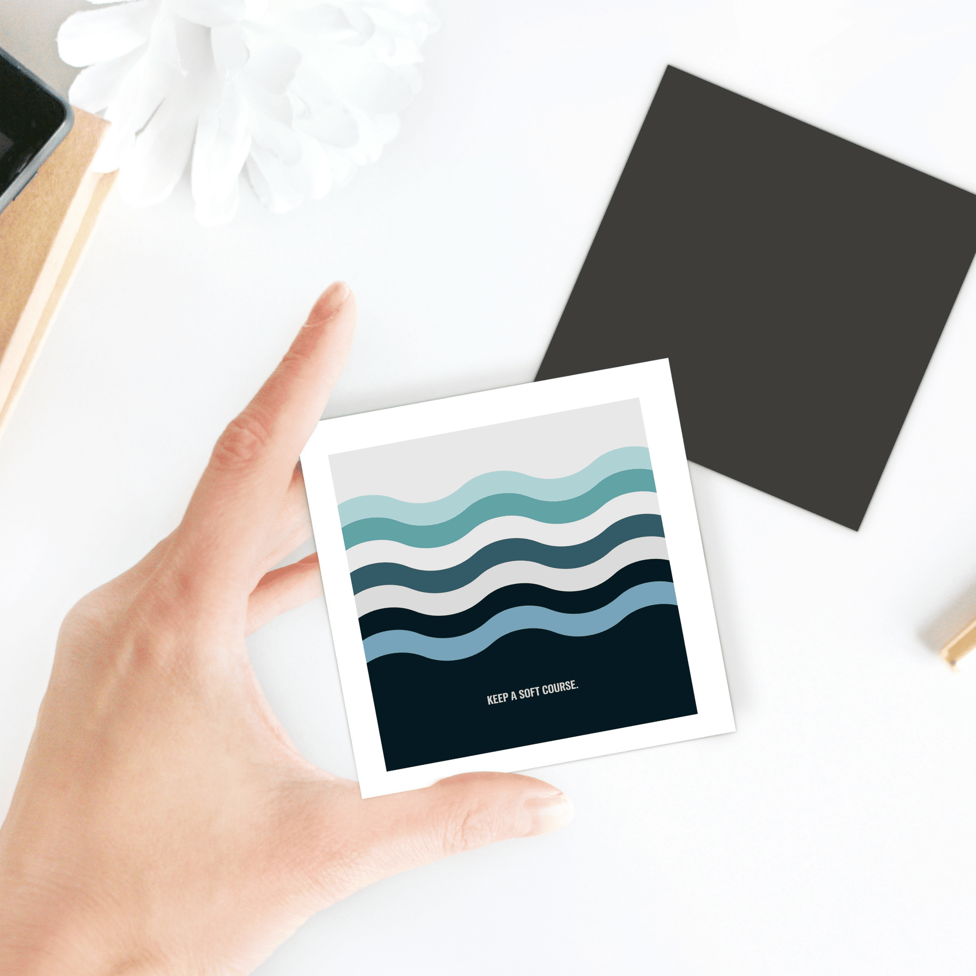 A hand holds Mirabilia Boutique’s “Keep a Soft Course Magnet – Minimalist Ocean-Inspired Design” over a white flower, black square paper, and part of a notebook—perfect for lovers of ocean vibes or minimalist decor.