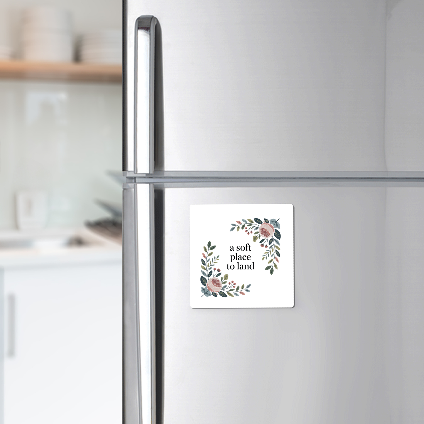 A silver fridge displays the Mirabilia Boutique "A Soft Place to Land Floral Magnet," a square cottagecore piece with a floral wreath and uplifting words. Blurred shelves and kitchen counter appear in the background.