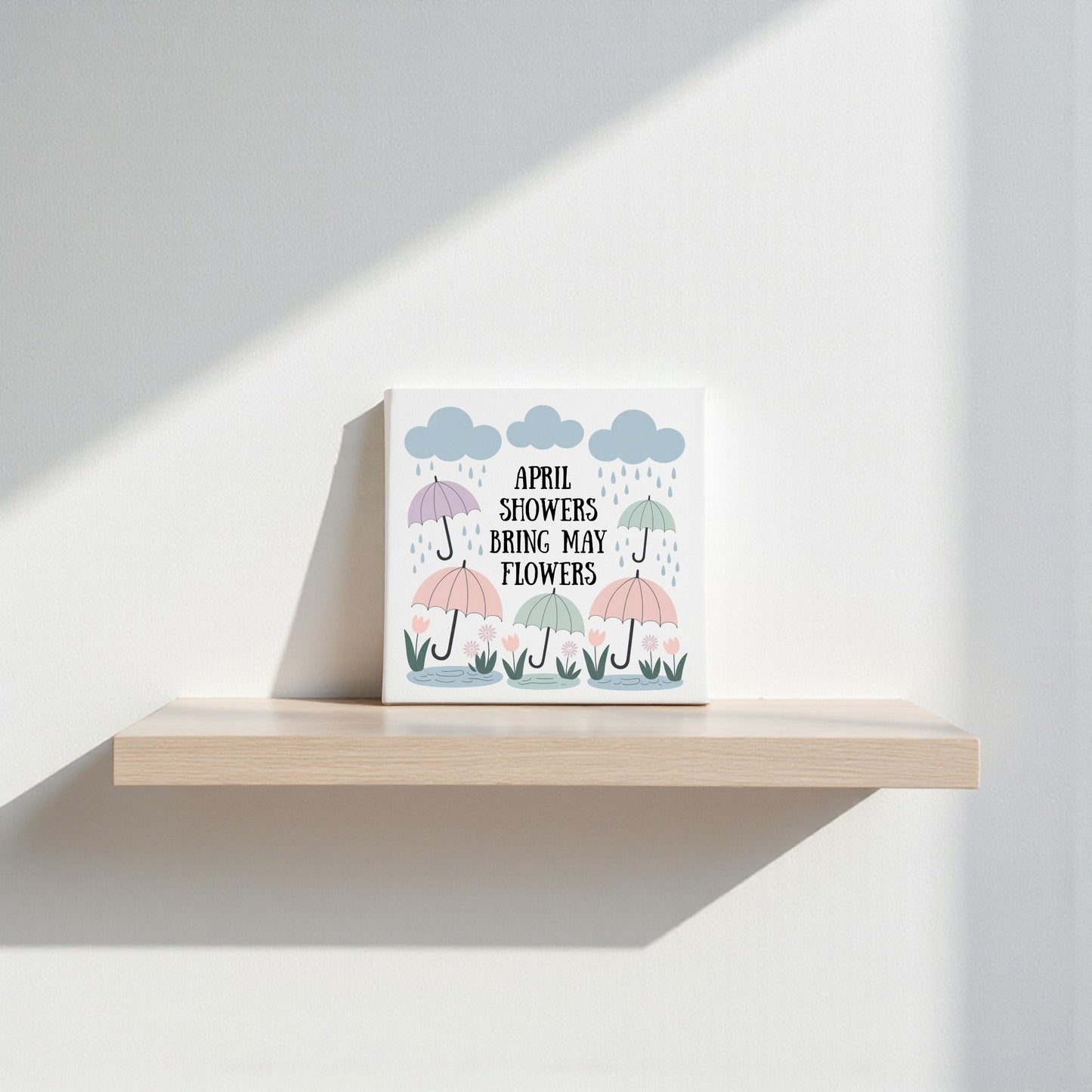 The April Showers Bring May Flowers Canvas Art by Mirabilia Boutique features pastel umbrellas and blooms under clouds, with its title at the center—charming spring wall decor ideal for nurseries or shelves.