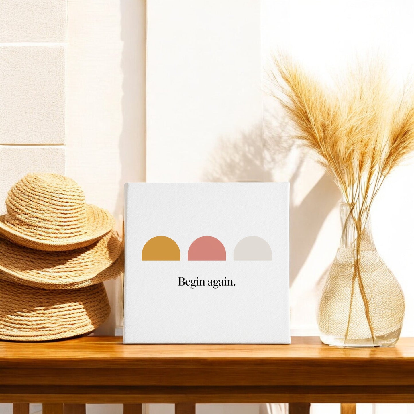 A wooden table with two straw hats, a glass vase of dried wheat, and Mirabilia Boutique’s "Begin Again Canvas – Soft Summer Affirmation Wall Art" featuring colored semi-circles, all basking in sunrise-inspired decor.