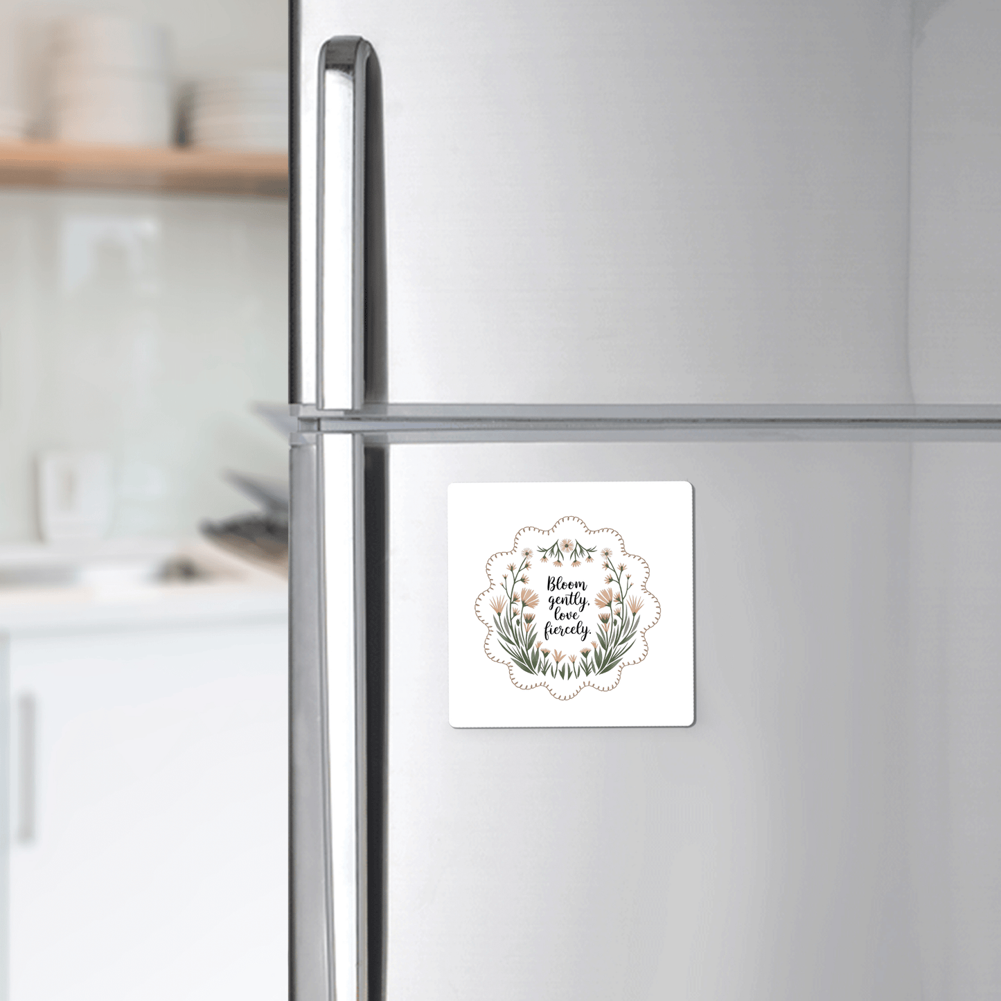 A silver refrigerator displays the Mirabilia Boutique Bloom Gently Love Fiercely Floral Magnet, a square botanical magnet featuring uplifting artwork and quote, adding charm to a modern kitchen.