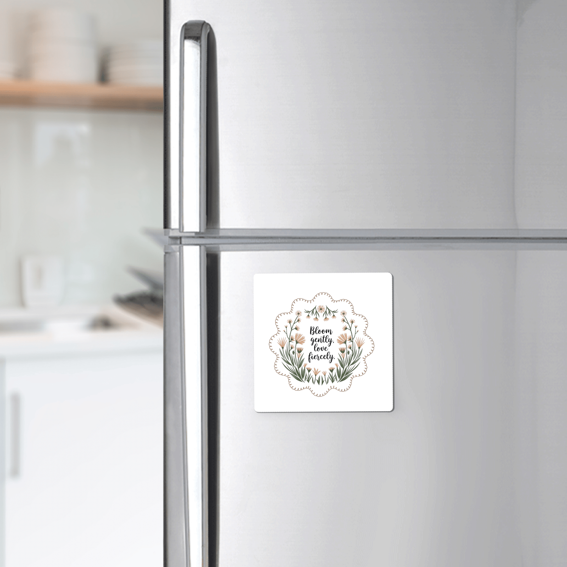 A silver refrigerator displays the Mirabilia Boutique Bloom Gently Love Fiercely Floral Magnet, a square botanical magnet featuring uplifting artwork and quote, adding charm to a modern kitchen.