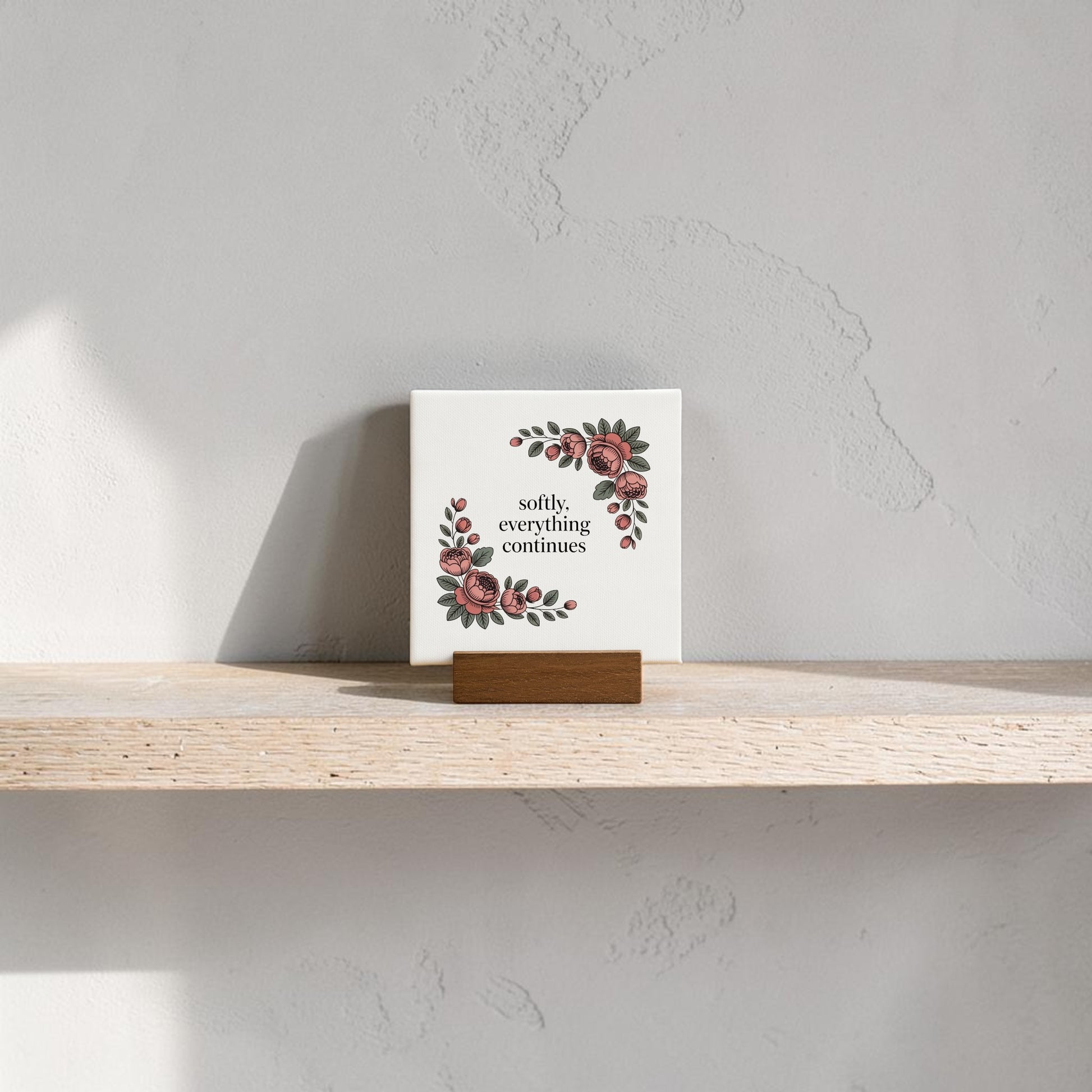 The Mirabilia Boutique Softly, Everything Continues Mini Sign, featuring floral designs and a wooden holder, sits on a shelf against a beige wall—perfect as a decorative accent or thoughtful gift.