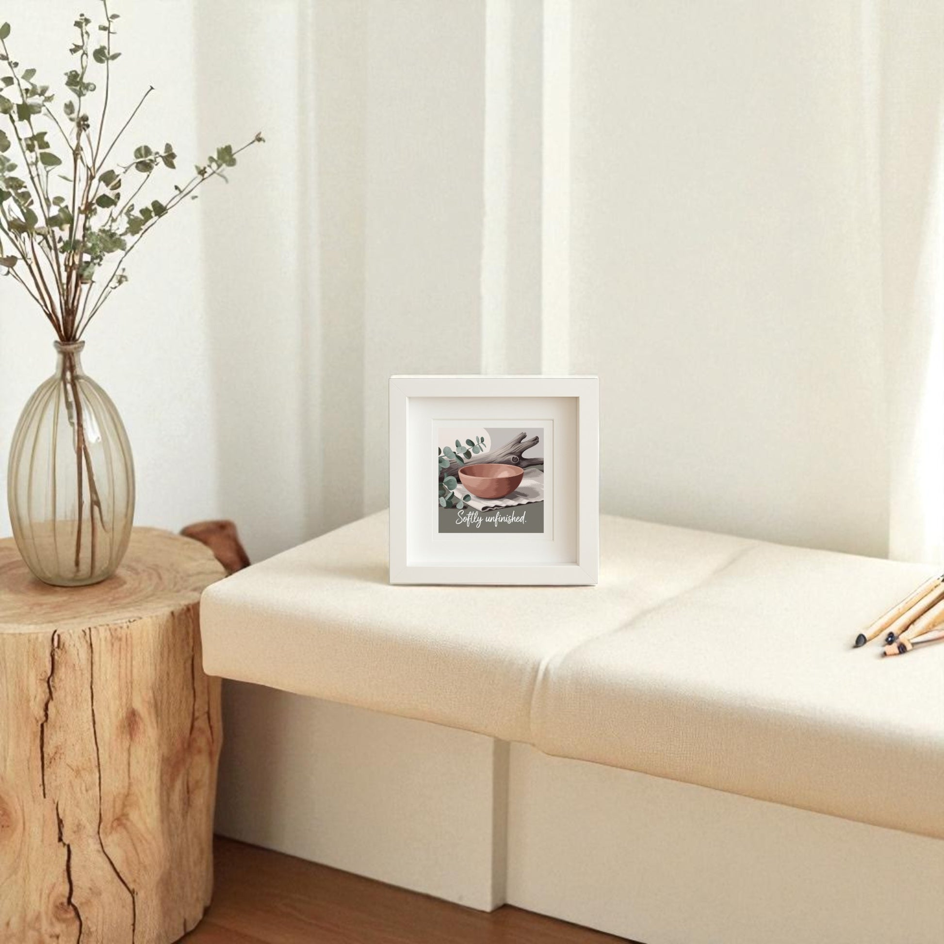 The Softly Unfinished Shadowbox by Mirabilia Boutique rests on a beige cushioned bench beside a rustic stool with greenery, showcasing modern boho style against a sunlit white wall.