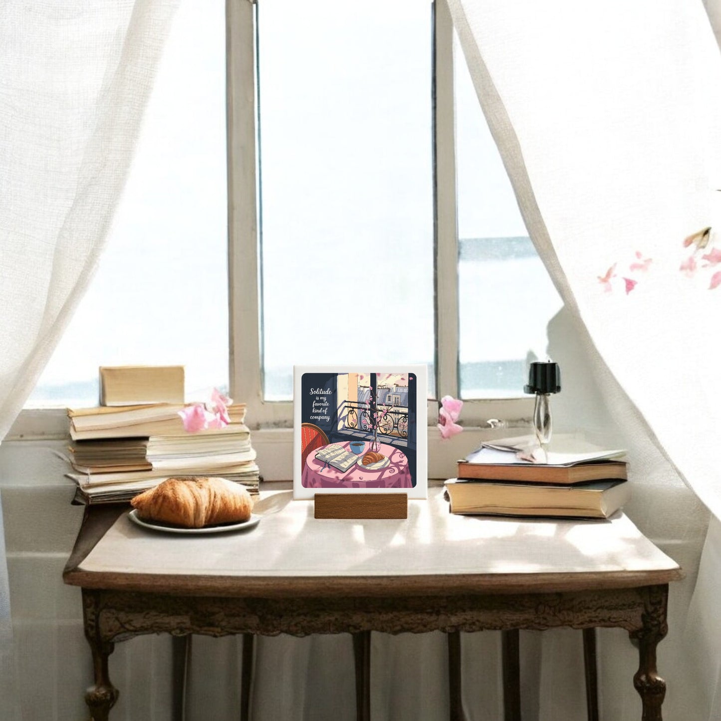 The Solitude Mini Sign – Parisian Morning Still Life by Mirabilia Boutique features a cozy scene with a croissant, books, a black vase with a single flower, and an open book on a sunlit table styled with Parisian balcony decor and sheer curtains.
