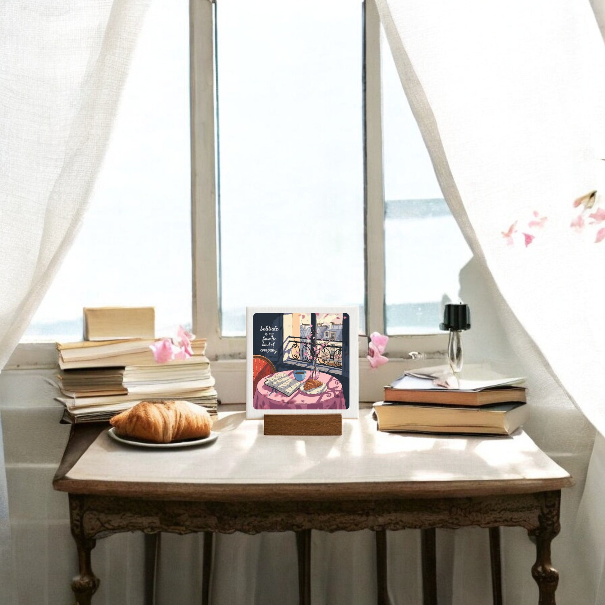 The Solitude Mini Sign – Parisian Morning Still Life by Mirabilia Boutique features a cozy scene with a croissant, books, a black vase with a single flower, and an open book on a sunlit table styled with Parisian balcony decor and sheer curtains.