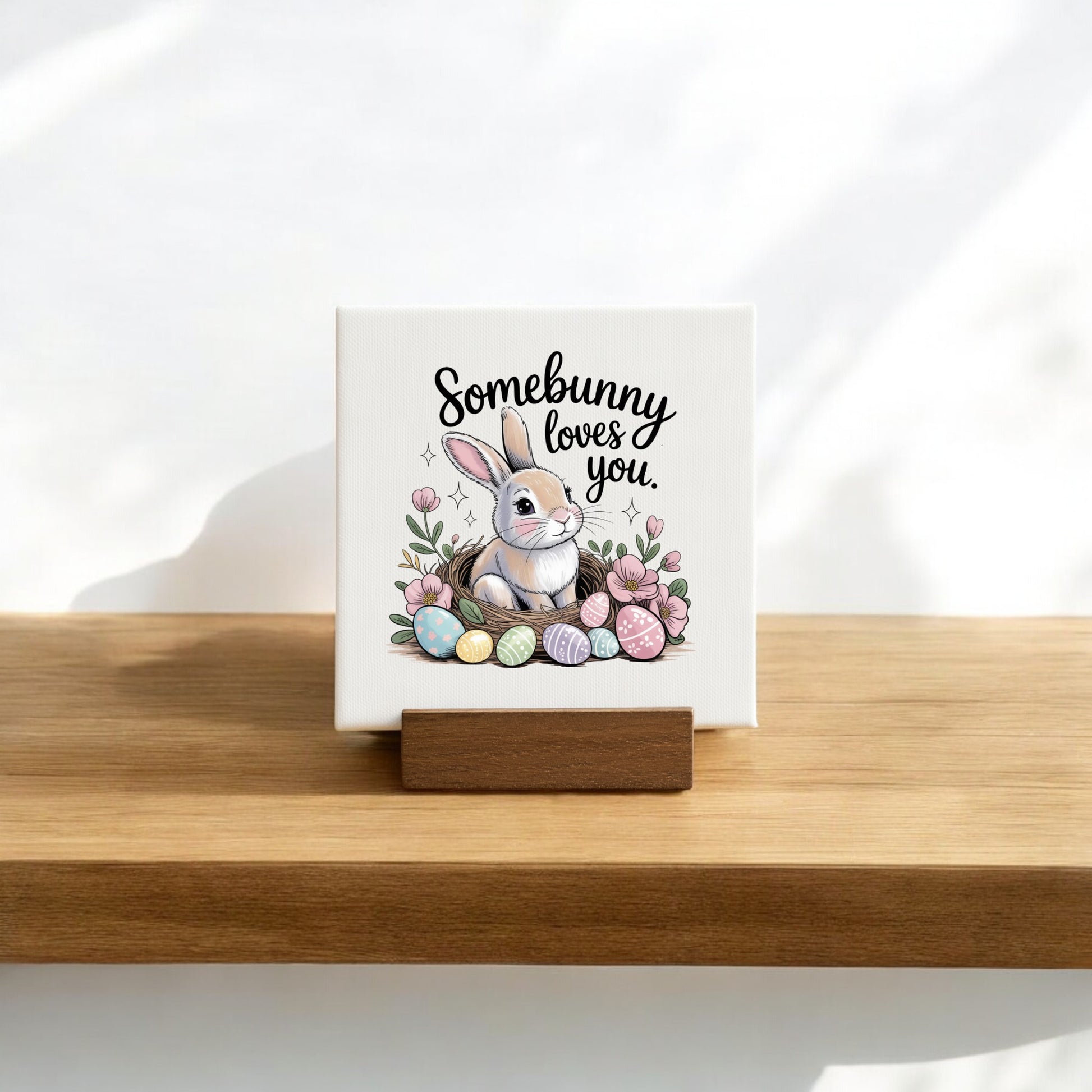 The Mirabilia Boutique Somebunny Loves You Mini Canvas (4x4) on a wood stand features a white bunny with pastel eggs, flowers, and sweet text—perfect for spring decor or as an Easter basket filler.