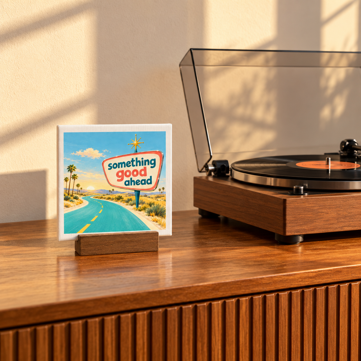 A Something Good Ahead Mini Canvas Sign | Chrome & Starlight featuring mid-century artwork of a road and palm trees, displayed as desk decor next to a record player.