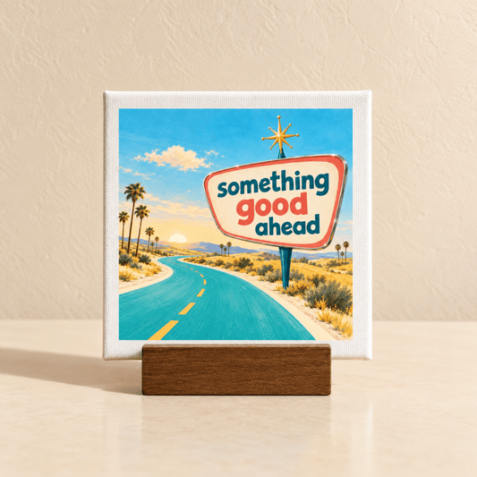 A Something Good Ahead Mini Canvas Sign | Chrome & Starlight sits on a wooden stand, depicting a mid-century style desert highway leading toward a sunset with a retro road sign that reads, “something good ahead.”.