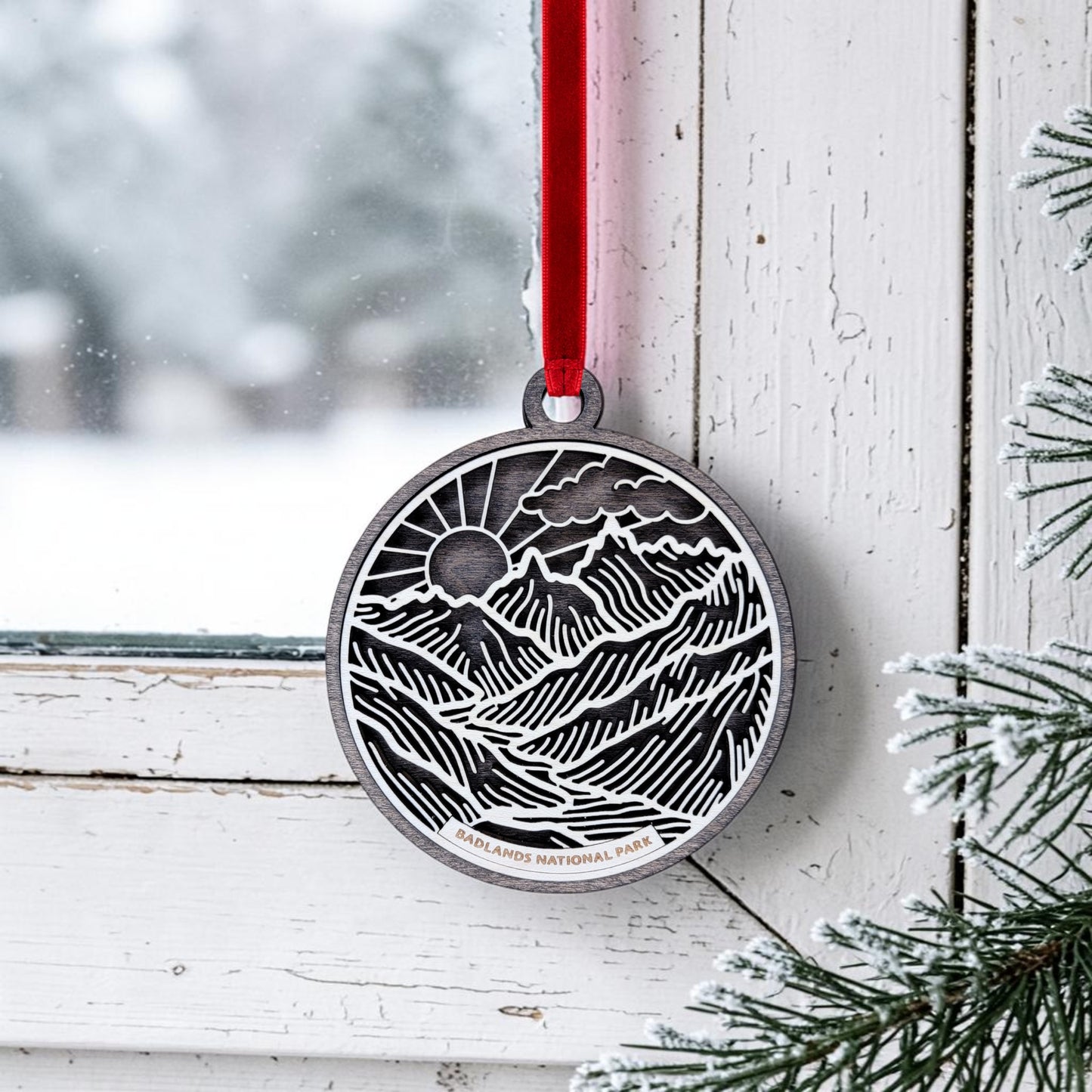 A round wooden keepsake, the Badlands National Park Line Art Ornament from Mirabilia Boutique, displays a mountain and sun design with "Badlands National Park" inscribed. It hangs by a red ribbon on a snowy window frame, with pine branches in the foreground enhancing its charm.