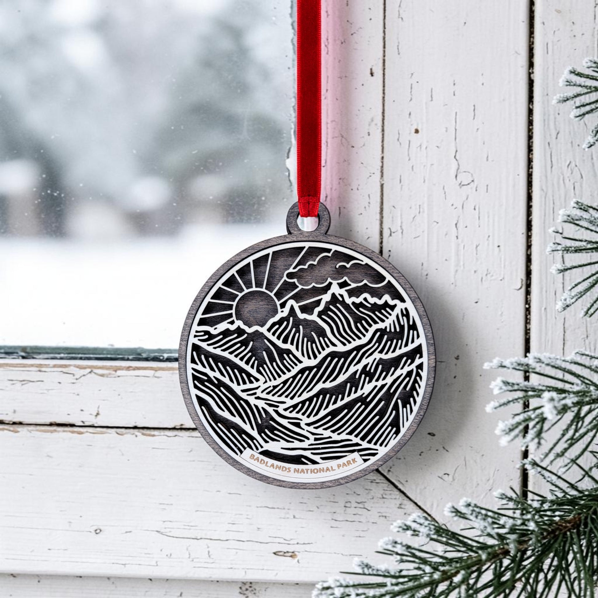 A round wooden keepsake, the Badlands National Park Line Art Ornament from Mirabilia Boutique, displays a mountain and sun design with "Badlands National Park" inscribed. It hangs by a red ribbon on a snowy window frame, with pine branches in the foreground enhancing its charm.