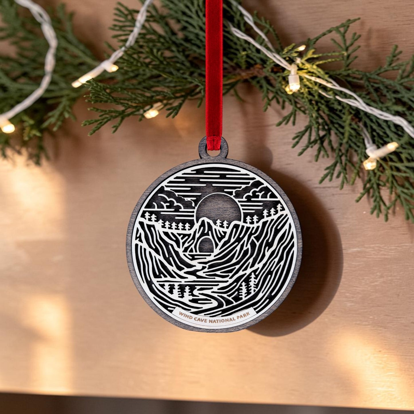 The Wind Cave National Park Line Art Christmas Ornament by Mirabilia Boutique is an oval, handcrafted wooden decoration showcasing a black and white illustration of mountains, trees, and a cave inspired by the Black Hills. Labeled “Wind Cave National Park,” it hangs from a red ribbon amidst greenery and string lights in the background.