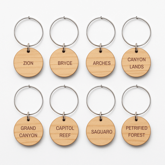 The Southwest National Park Wine Charms Set by Mirabilia Boutique includes eight round wooden keychains with metal rings, each engraved with a park name—an ideal rustic gift for adventurers and national park lovers.