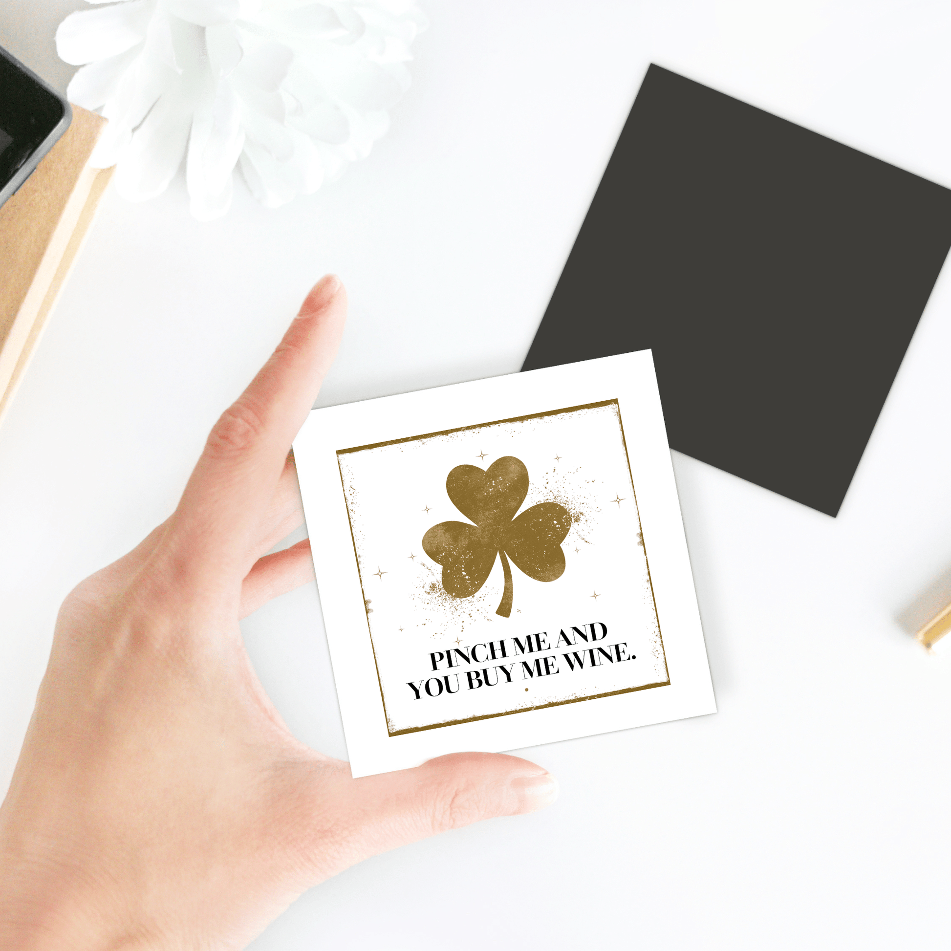 A hand holds the Mirabilia Boutique "Pinch Me and You Buy Me Wine Magnet" with a gold shamrock on white—fun St. Patrick’s Day or wine lover gift. Nearby: a black square card and white flower rest on a white surface.