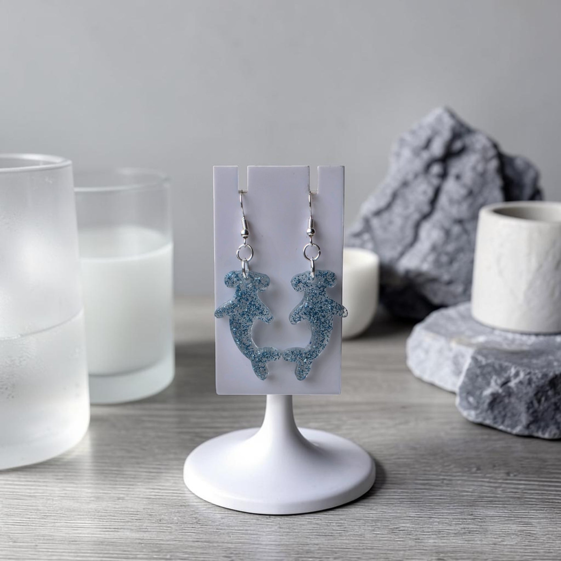 A pair of Blue Glitter Hammerhead Shark Earrings by Mirabilia Boutique hang on a white display stand, set on a wooden surface with candles, glass cups, and gray stones—ideal for fans of ocean-themed jewelry.