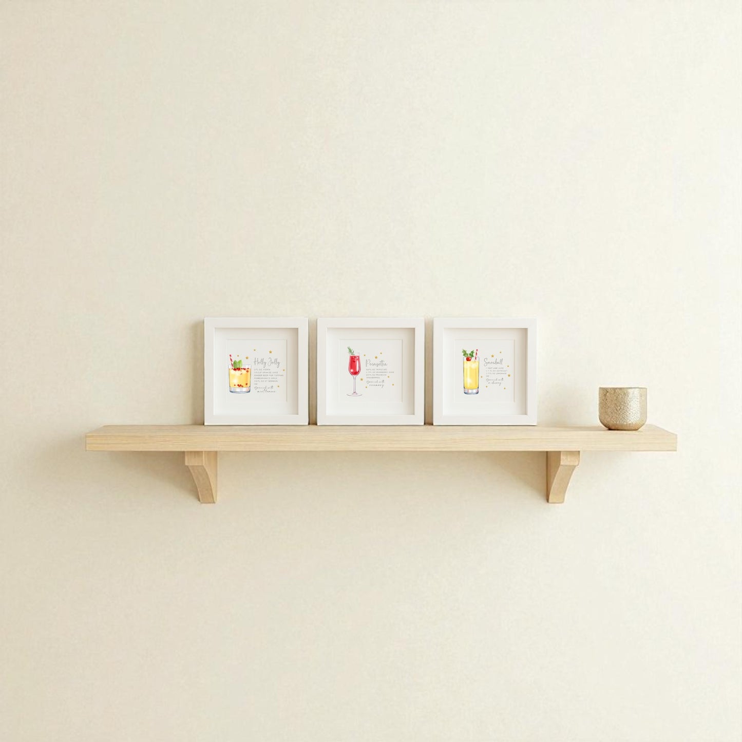 A light wooden wall shelf displays the Mirabilia Boutique Sparkly Holiday Cocktails 5x5 Shadowbox Trio festive bar art and a small beige textured cup, all set against a plain off-white wall.