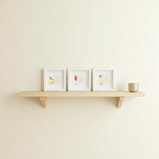 A light wooden wall shelf displays the Mirabilia Boutique Sparkly Holiday Cocktails 5x5 Shadowbox Trio festive bar art and a small beige textured cup, all set against a plain off-white wall.