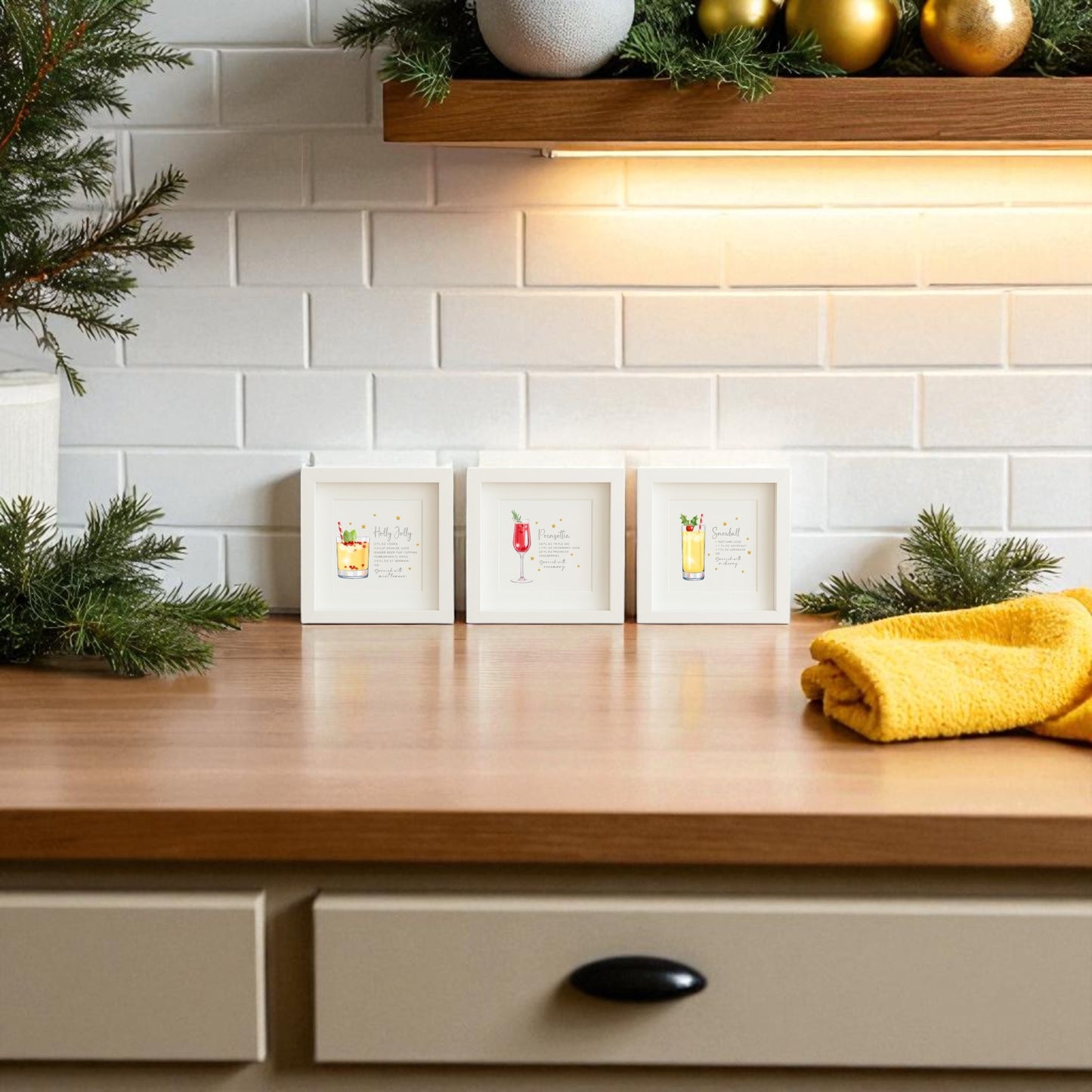 A kitchen countertop features a yellow cloth, festive greenery, and Mirabilia Boutique’s Sparkly Holiday Cocktails 5x5 Shadowbox Trio art. White subway tile backsplash and a shelf with gold ornaments complete the festive look.