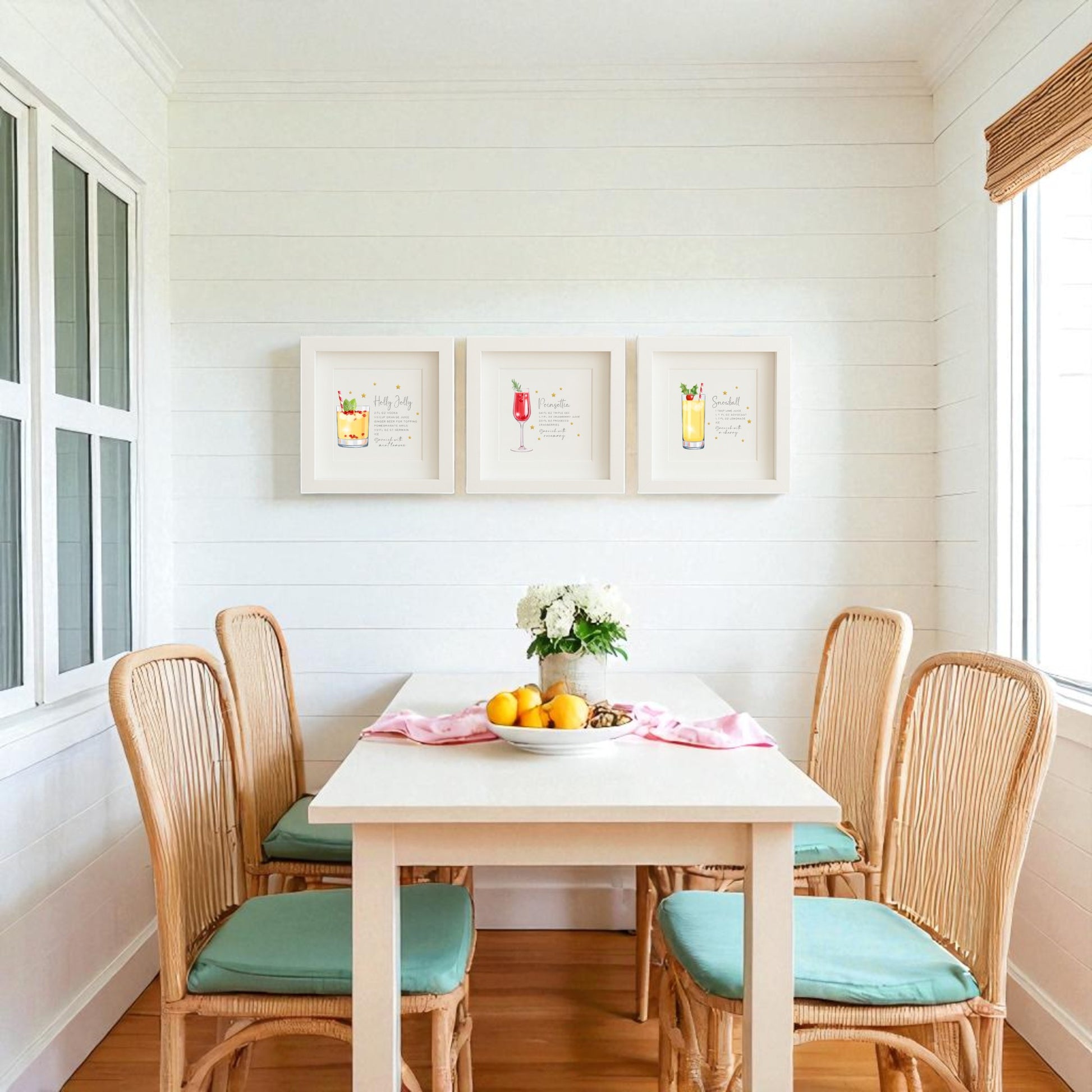 A bright dining area with a white table for four, wicker chairs with blue cushions, lemons and flowers as decor, and Mirabilia Boutique’s Sparkly Holiday Cocktails 5x5 Shadowbox Trio festive bar art on the shiplap wall. Natural light streams in.