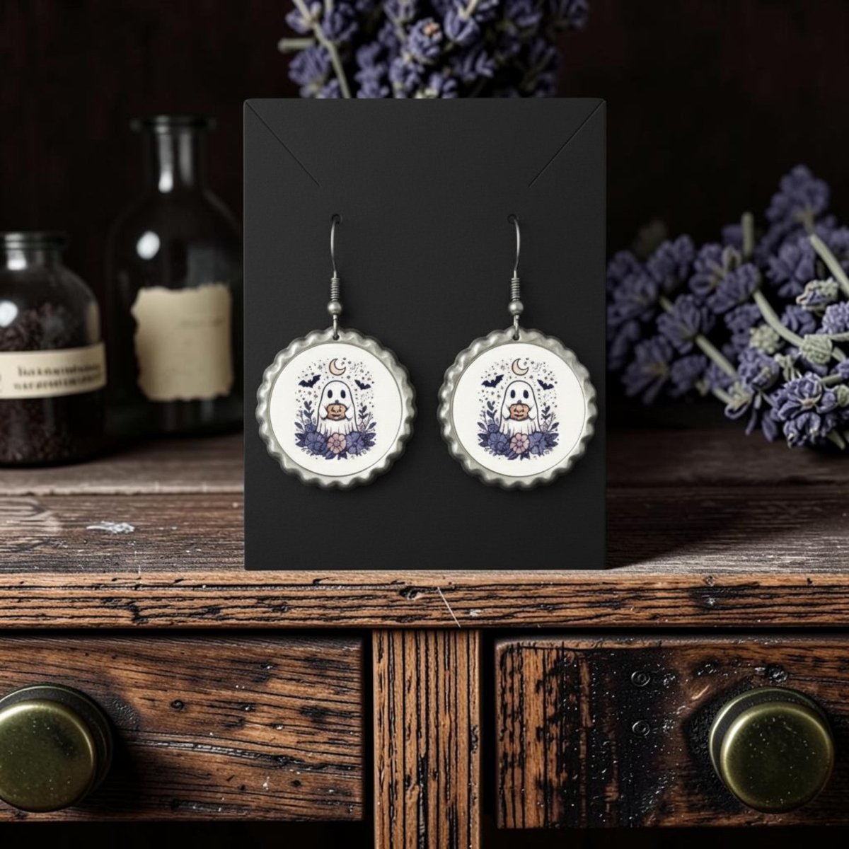 A pair of Crescent Moon Pumpkin Ghost Scalloped Earrings by Mirabilia Boutique, featuring bottlecap-shaped frames with ghost and floral designs, displayed on a black card atop a rustic wooden table with dried lavender and apothecary jars in the background.