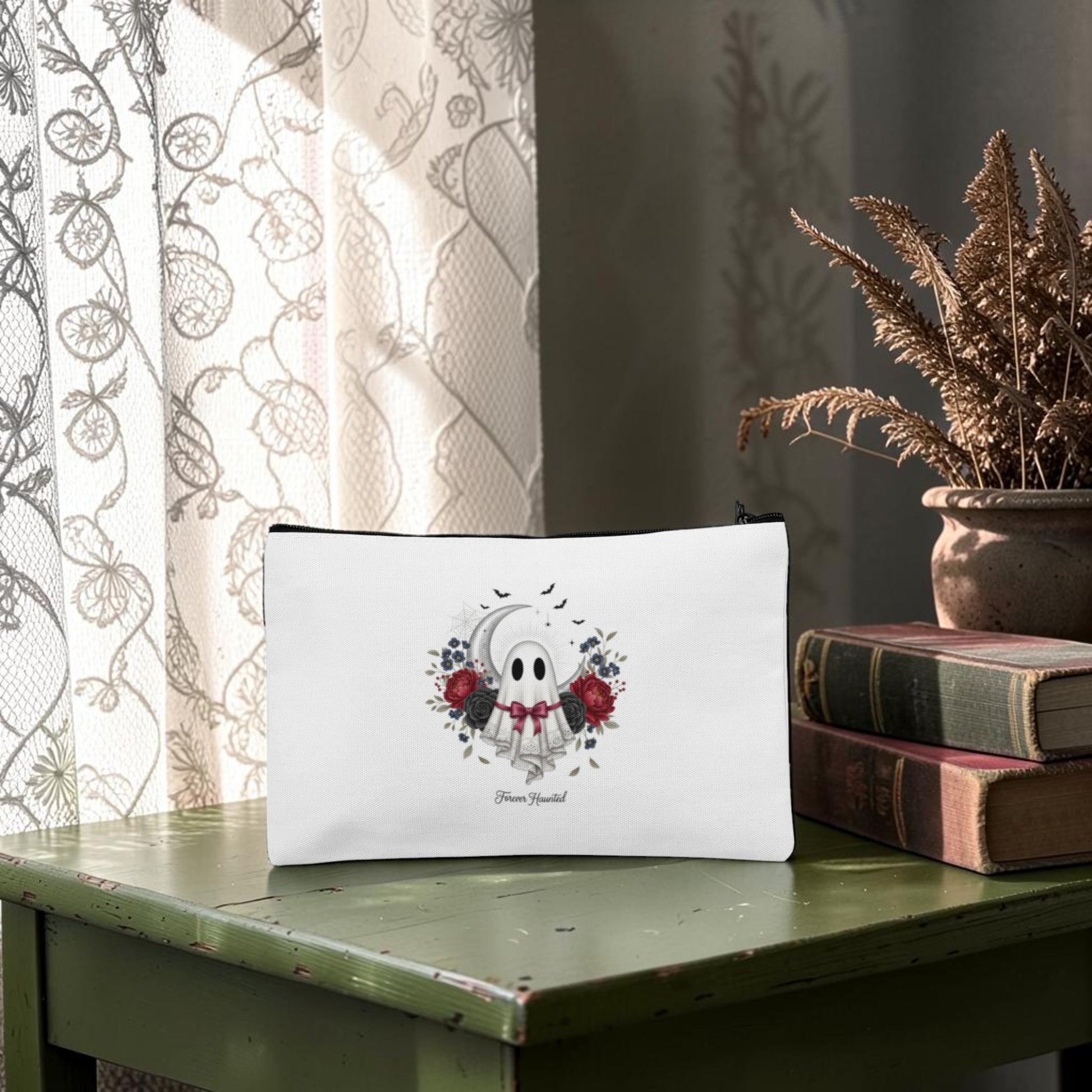 The Forever Haunted Makeup Bag by Mirabilia Boutique—a white pouch adorned with a charming ghost illustration and delicate floral details—sits on a green table next to stacked vintage books and a vase of dried grass by a lace-curtained window, making it the perfect gothic ghost pouch or spooky season accessory.