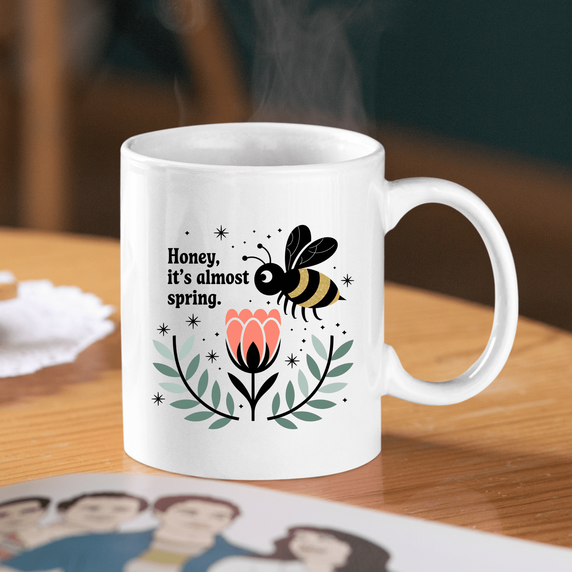 The Mirabilia Boutique Spring Bee Mug | Cute “Honey, It’s Almost Spring” Coffee Cup sits on a wooden table, decorated with a bee above a flower and green leaves. Steam rises from the mug, with a family photo partially visible nearby.