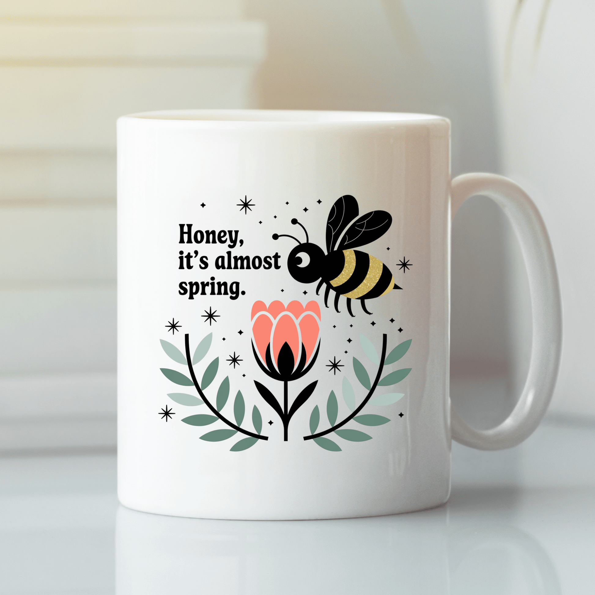 The Spring Bee Mug by Mirabilia Boutique features a bee above a pink flower with green leaves and the phrase “Honey, It’s Almost Spring,” making this cute coffee cup perfect for welcoming the season.