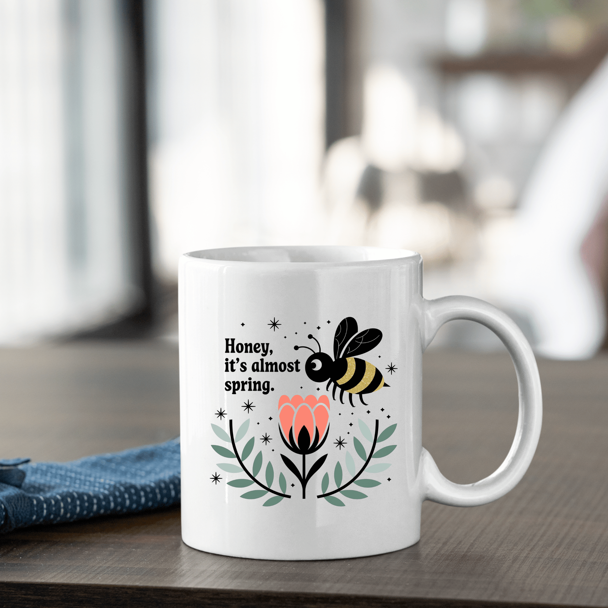 The Mirabilia Boutique Spring Bee Mug features a cute bee, pink flower, green leaves, and “Honey, It’s Almost Spring” text—perfect for coffee. This charming mug adds a touch of spring to any table.