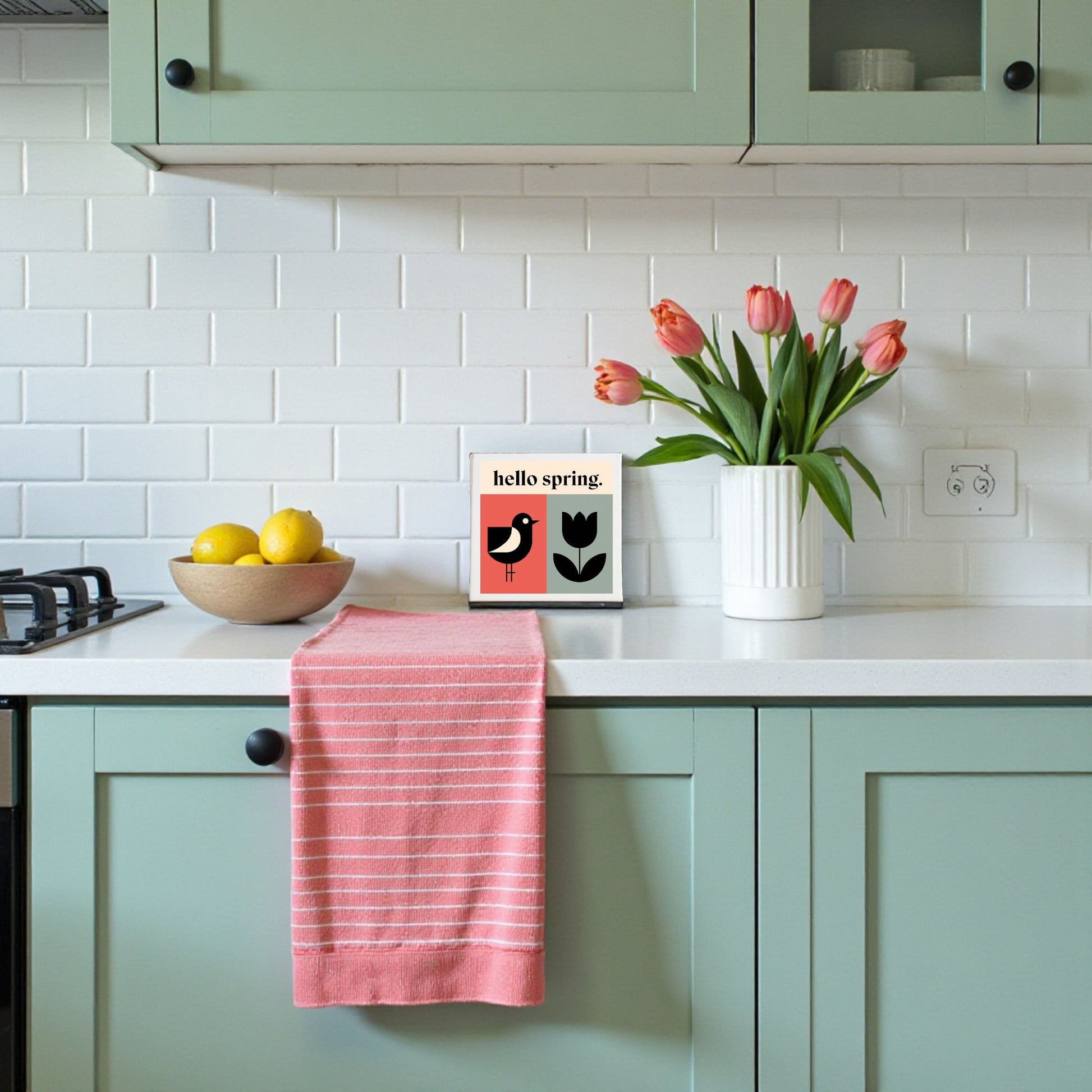 A kitchen with mint green cabinets, a white countertop, a bowl of lemons, a pink striped dish towel, and cheerful decor like Mirabilia Boutique’s Hello Spring Canvas – Bird & Tulip Wall Art, plus pink tulips against white subway tile.