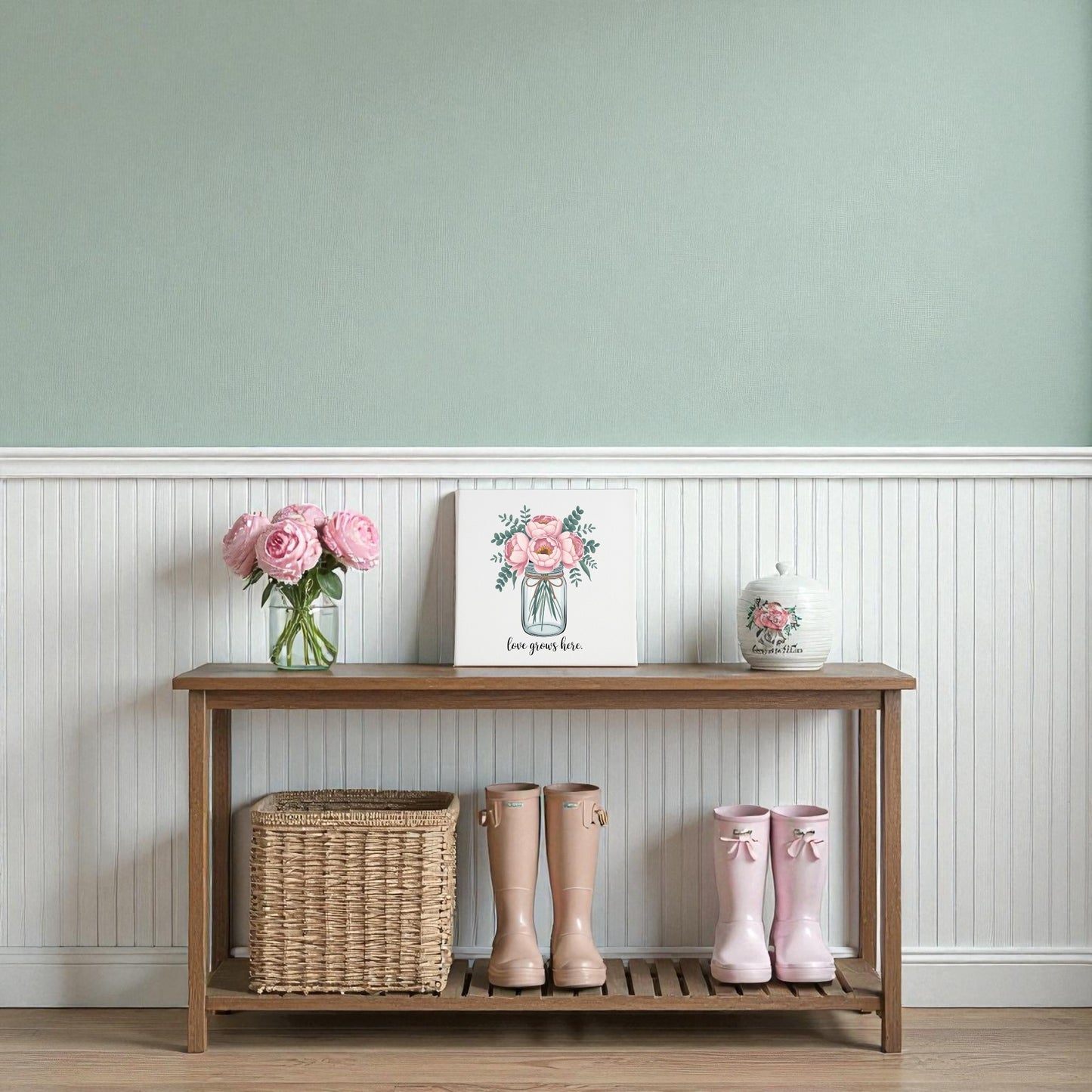 A wooden bench holds a woven basket, two pairs of pink rain boots, a vase of pink flowers, and the "Love Grows Here Floral Canvas Wall Art" by Mirabilia Boutique, all set against a pale green wall with white wainscoting.