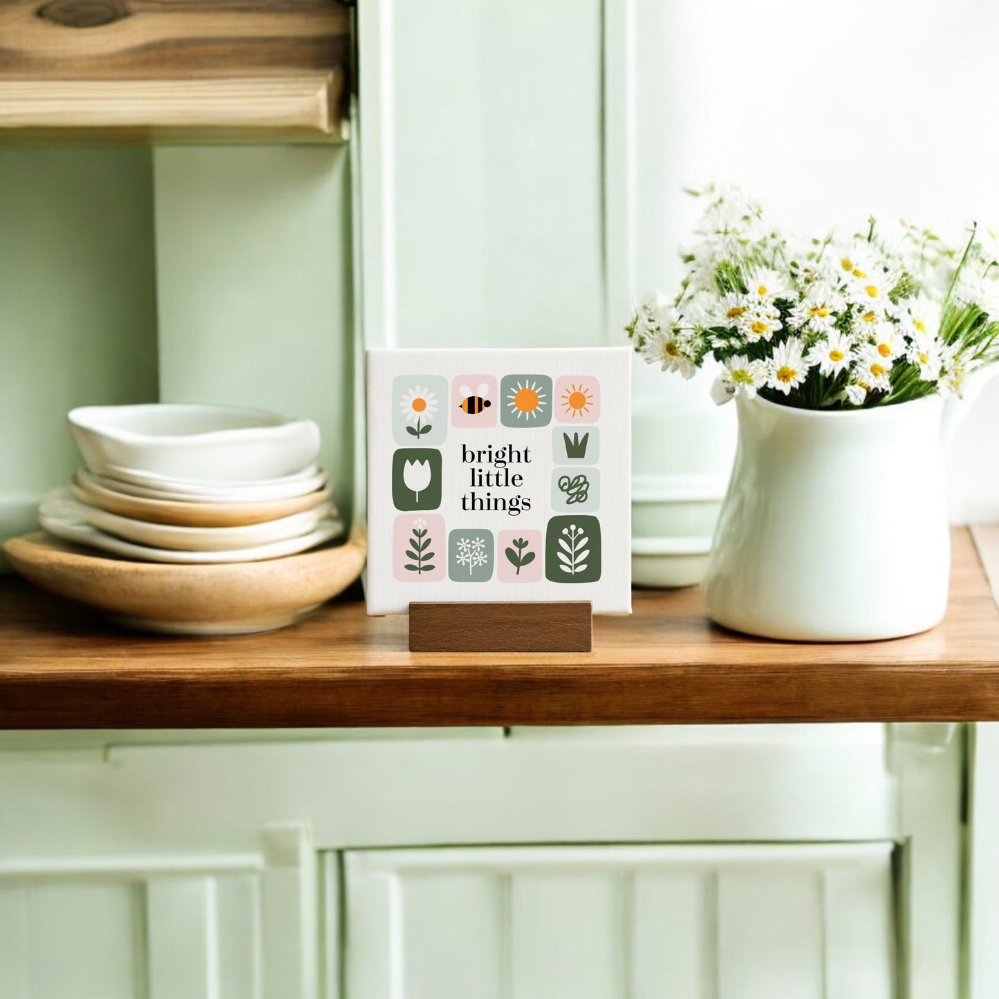 The Bright Little Things Mini Canvas Sign by Mirabilia Boutique, featuring floral decor, stands on a wooden shelf beside stacked dishes and a white jug of daisies, with light green cabinets in the background.