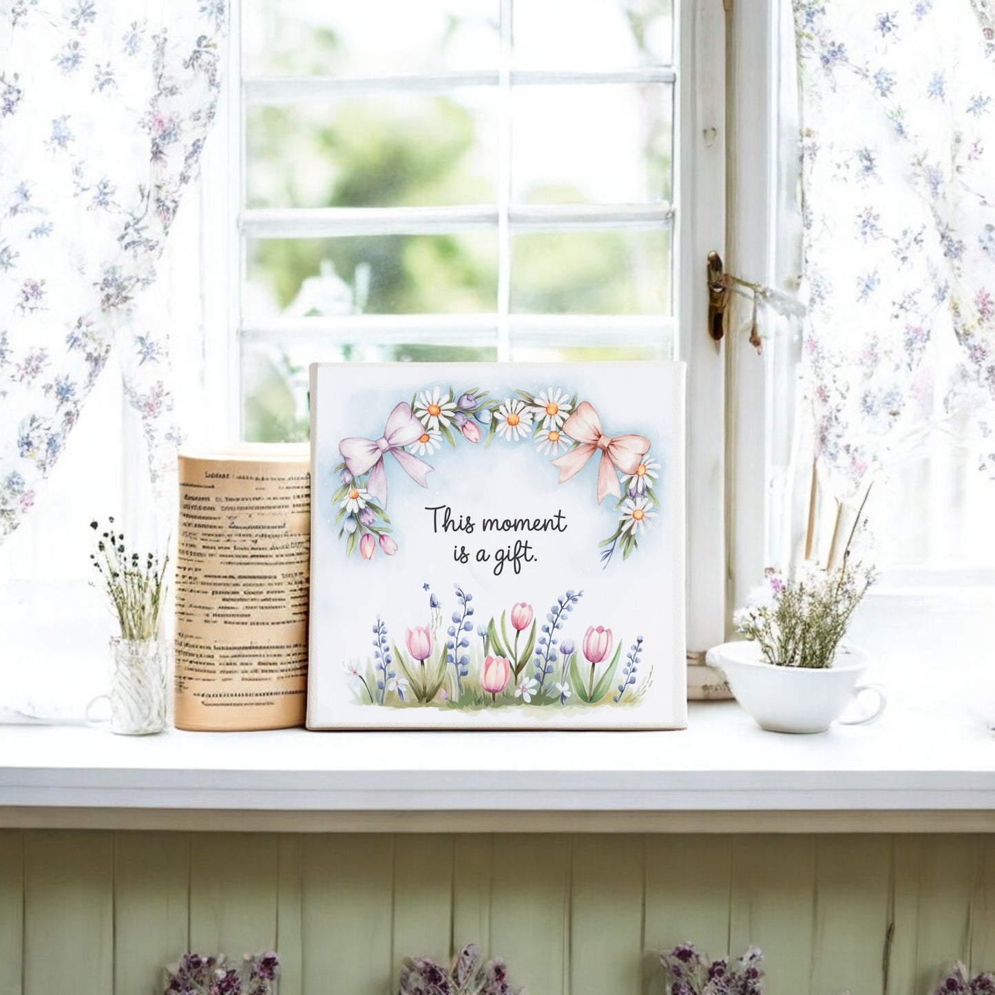 A bright windowsill with floral curtains displays a cup, a book, and the This Moment Is a Gift Canvas – 8x8 Spring Floral Art by Mirabilia Boutique. Jars of greenery nearby complete the perfect slow living decor.
