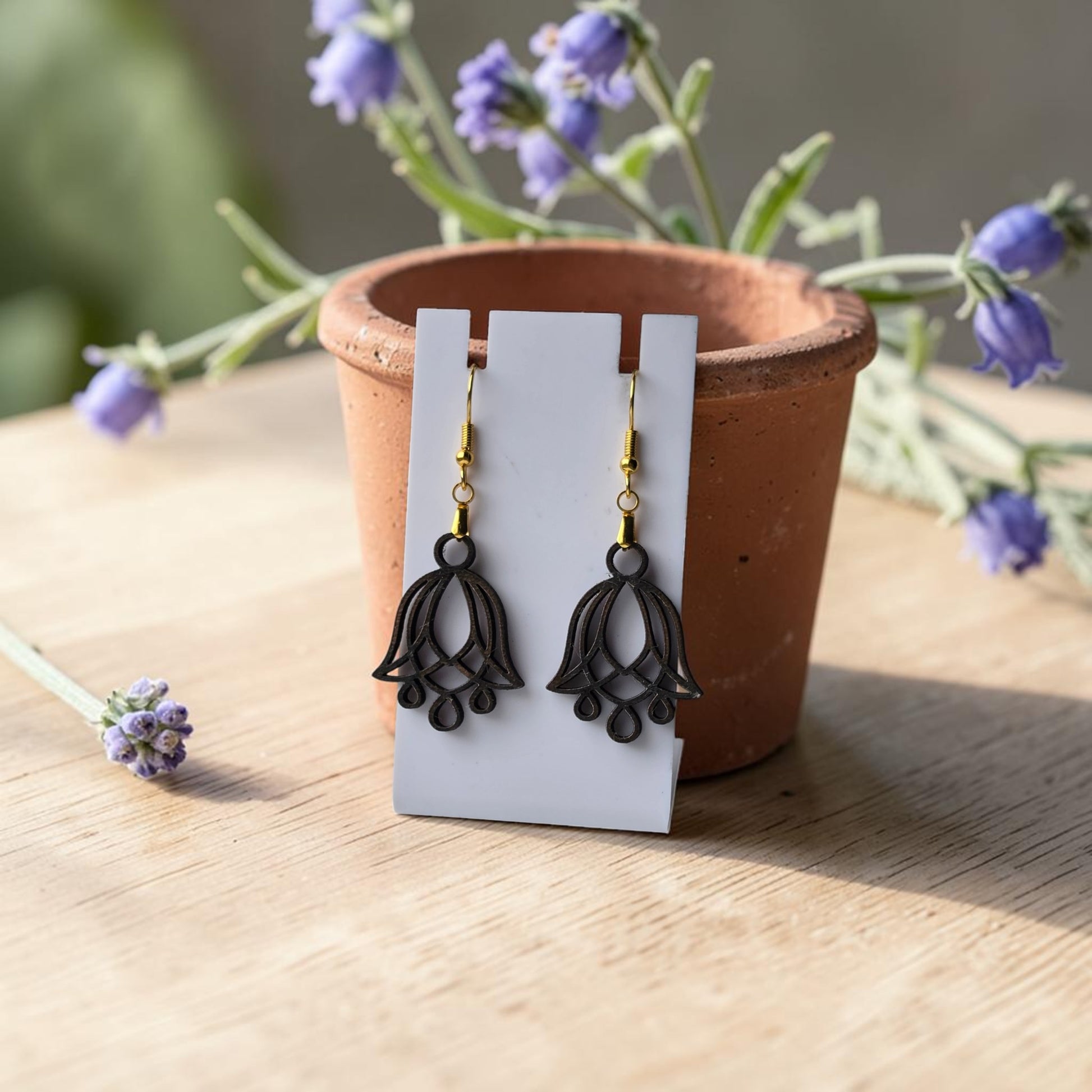 Bluebell Flower Earrings by Mirabilia Boutique, crafted from lasercut walnut wood, hang on a white card propped against a terracotta pot with purple flowers, all placed on a light wooden surface.