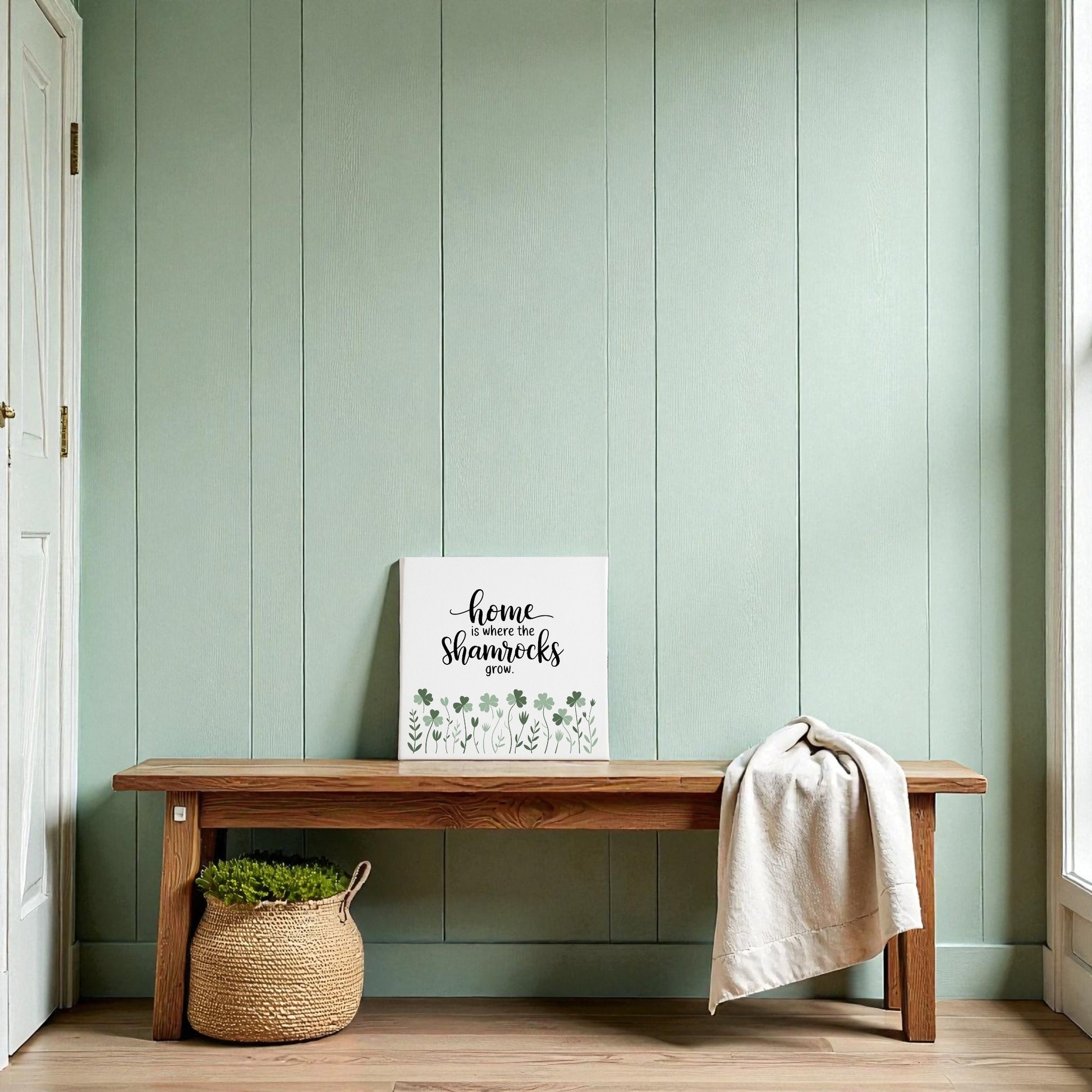 A wooden bench sits against a light green wall, topped with Mirabilia Boutique’s “Home Is Where the Shamrocks Grow” canvas and a cozy throw—perfect for adding Irish decor charm. A basket with a green plant rests nearby.