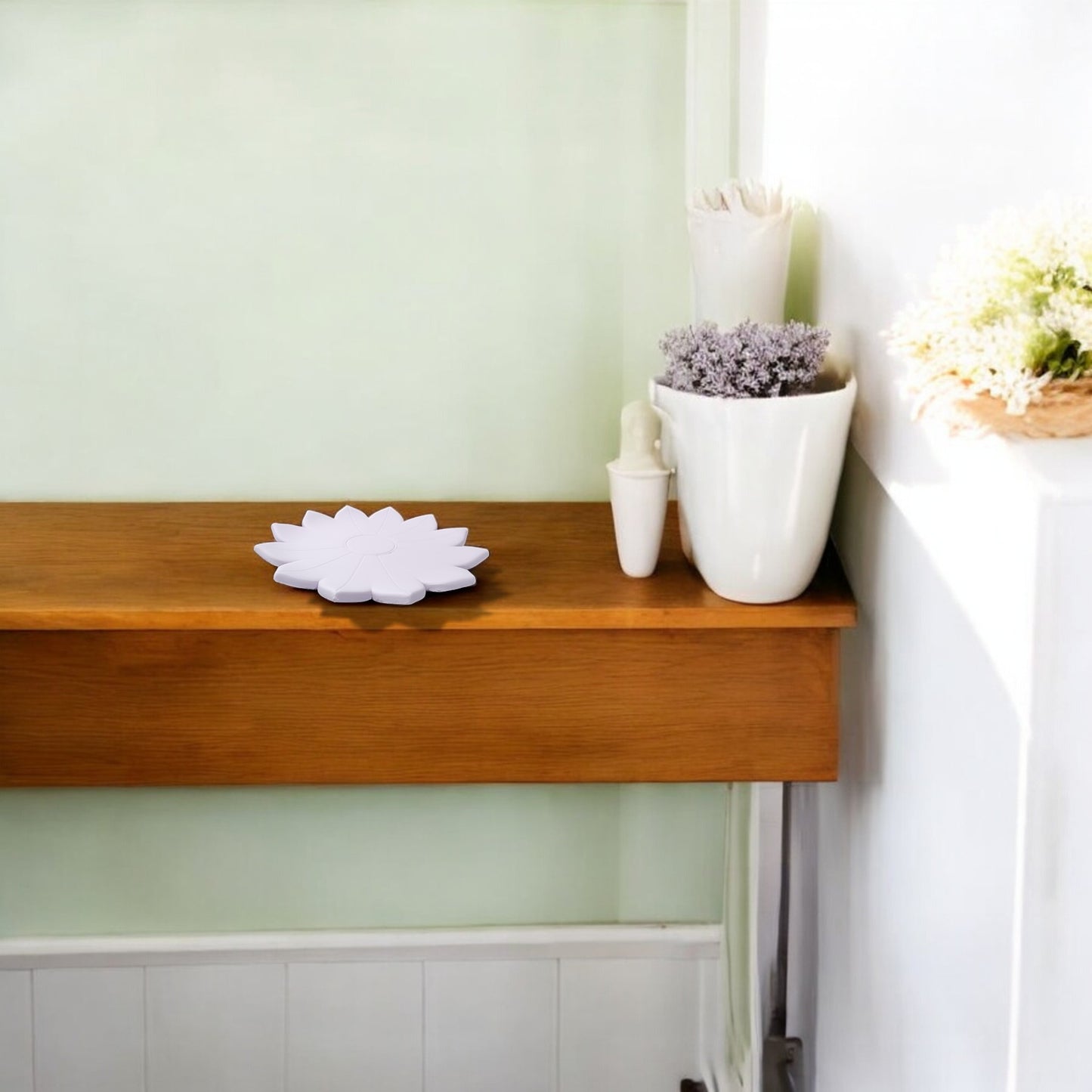 A wooden shelf displays the Mirabilia Boutique Spring Bloom Tray—a white, lotus-shaped accent—alongside a white vase with purple flowers, a smaller white vase, and a white pot of green and white blooms against pale green and white walls.