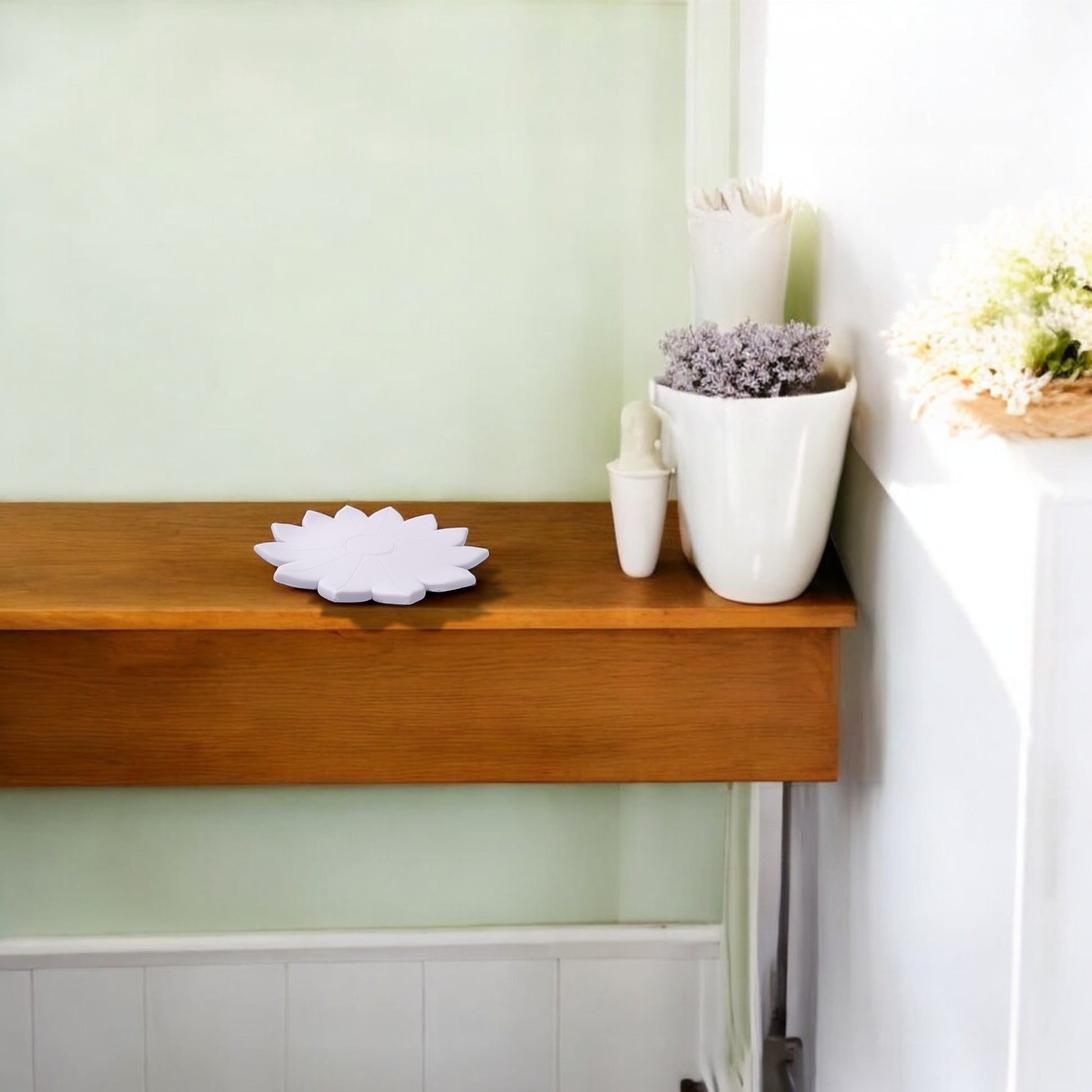 A wooden shelf displays the Mirabilia Boutique Spring Bloom Tray—a white, lotus-shaped accent—alongside a white vase with purple flowers, a smaller white vase, and a white pot of green and white blooms against pale green and white walls.