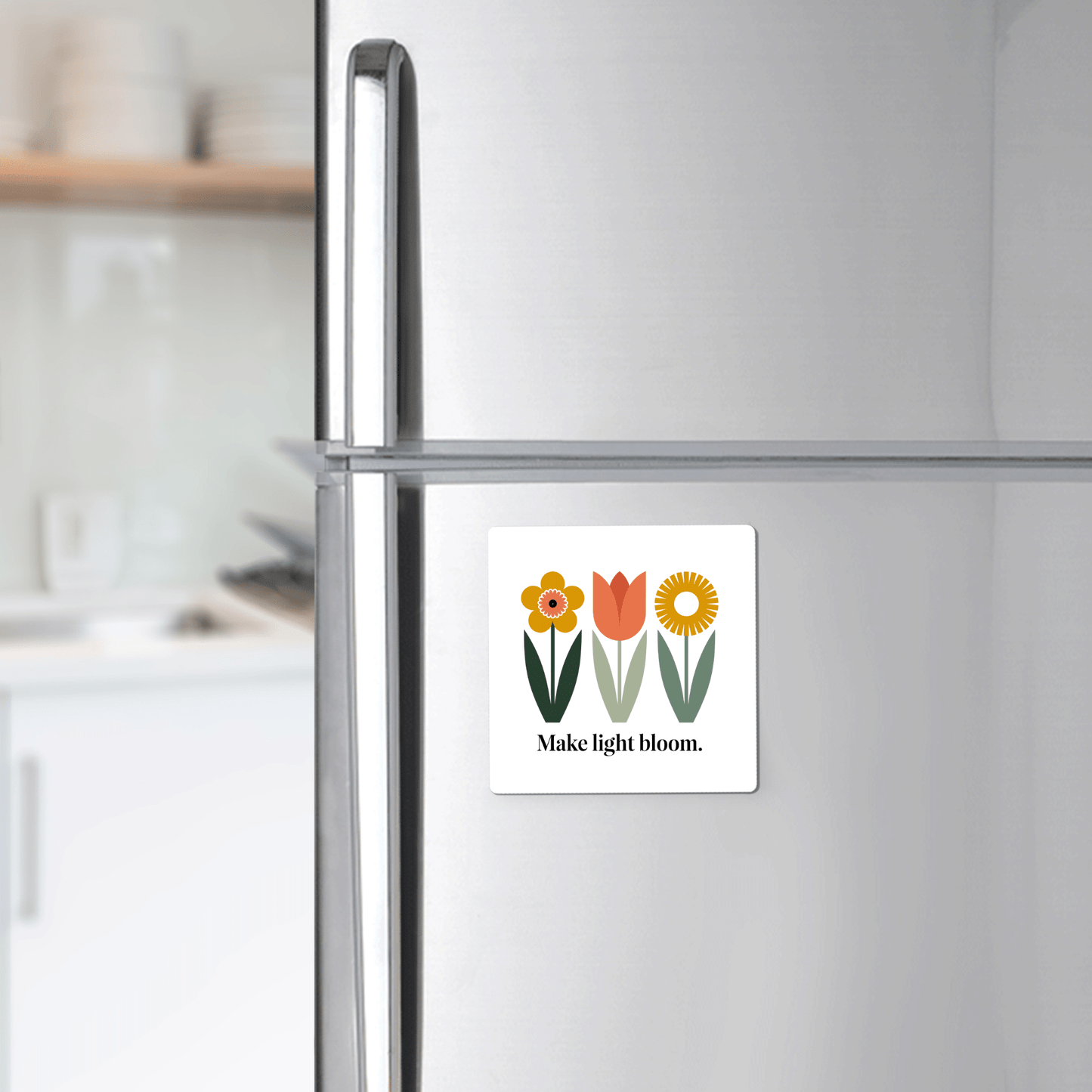A silver refrigerator showcases the Mirabilia Boutique “Make Light Bloom Magnet – Spring Floral Inspiration,” featuring three illustrated flowers and uplifting text. A softly blurred kitchen in the background enhances the fresh spring vibe.