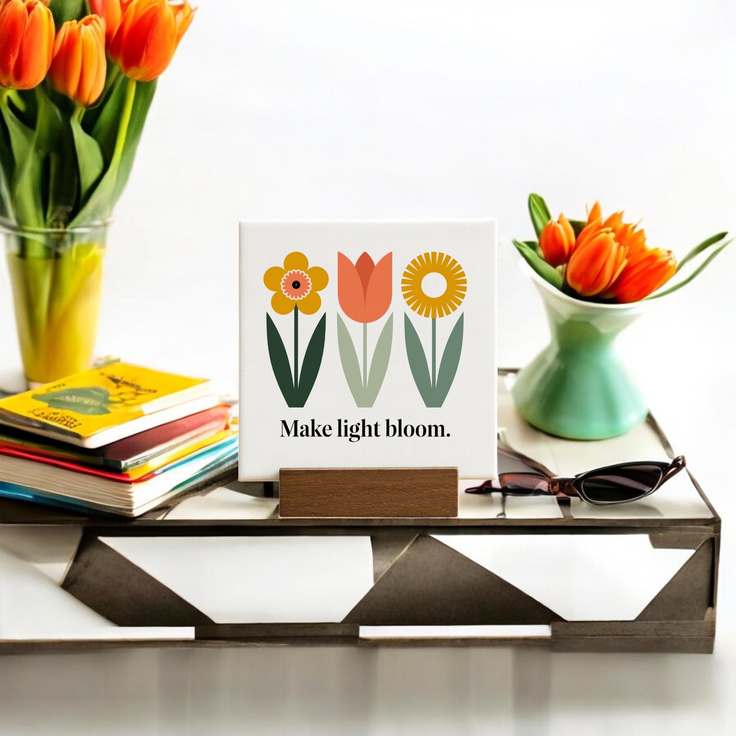 The Make Light Bloom Mini Canvas Sign with Wood Base by Mirabilia Boutique features three flower illustrations and an uplifting message, making it a perfect daily affirmation decor for your white table.