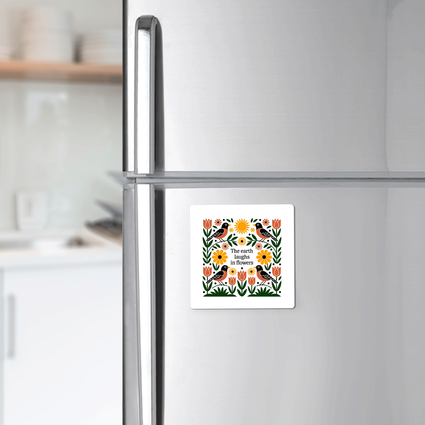 A silver refrigerator features the Mirabilia Boutique "The Earth Laughs in Flowers" Floral Magnet, decorated with flowers, birds, and a sun with the quote "The earth laughs in flowers." A white kitchen cabinet stands behind.