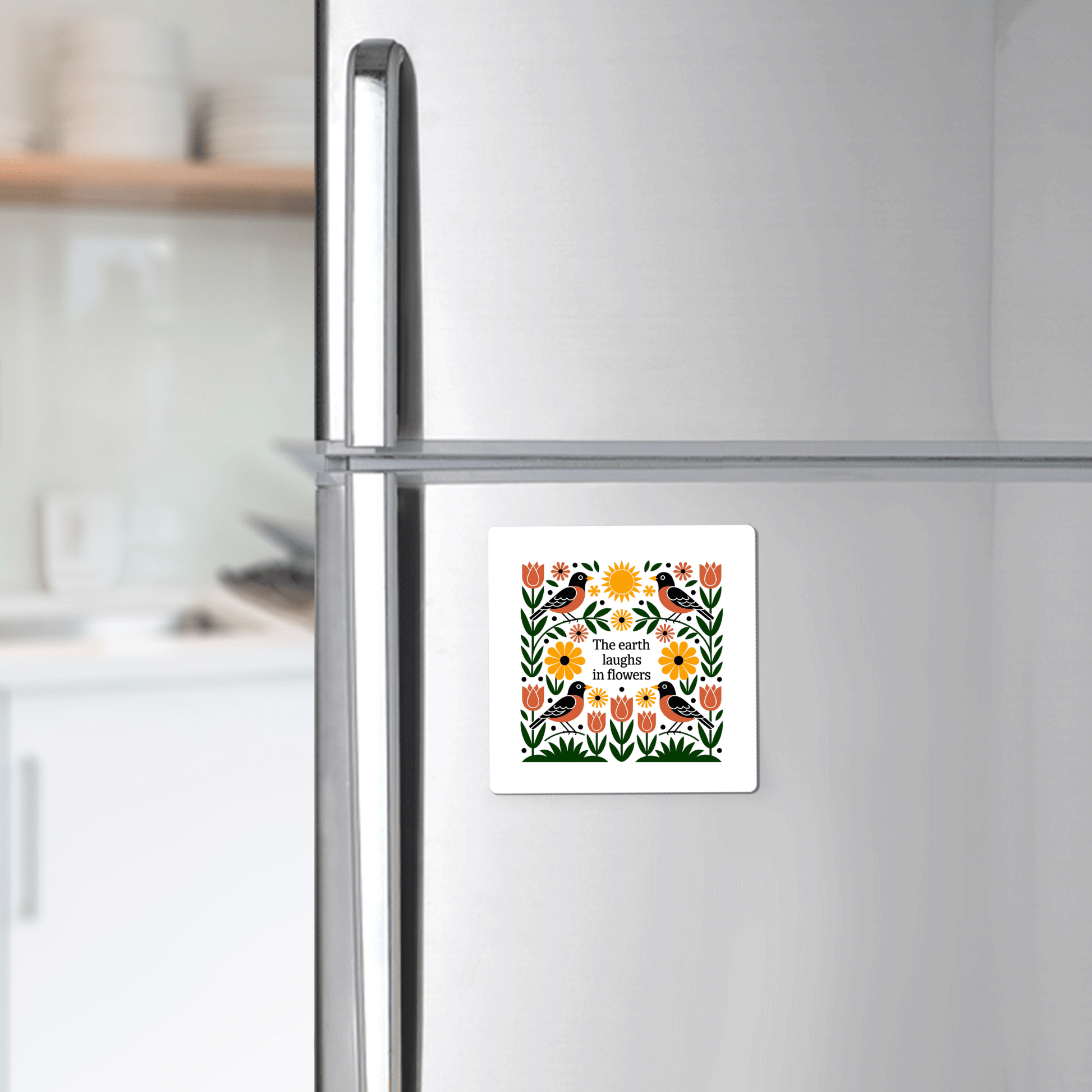 A silver refrigerator features the Mirabilia Boutique "The Earth Laughs in Flowers" Floral Magnet, decorated with flowers, birds, and a sun with the quote "The earth laughs in flowers." A white kitchen cabinet stands behind.