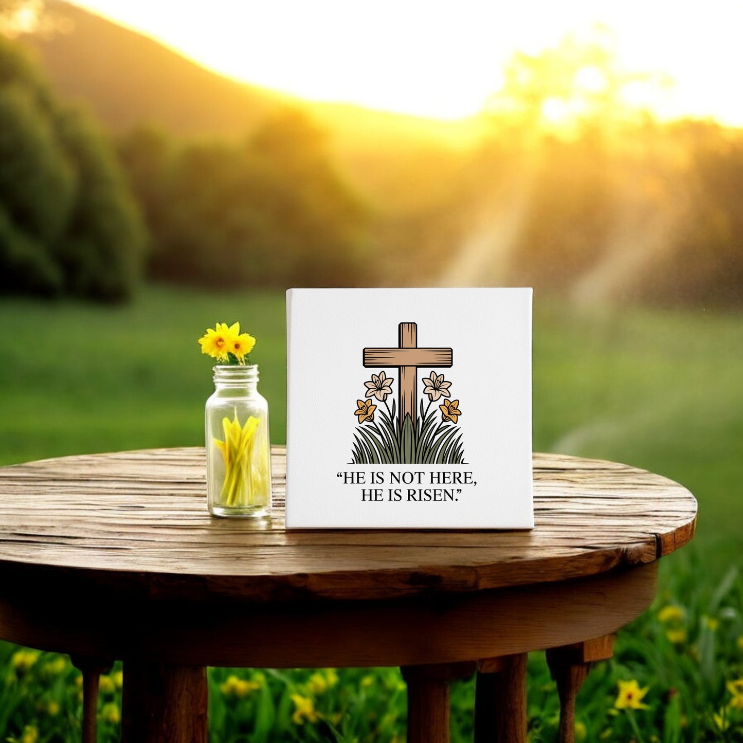 A small vase with yellow flowers and the Mirabilia Boutique He Is Risen Cross & Lily Canvas (8x8 Easter Art) sits on a wooden table outdoors, bathed in sunlight—perfect resurrection decor for your Easter celebration.