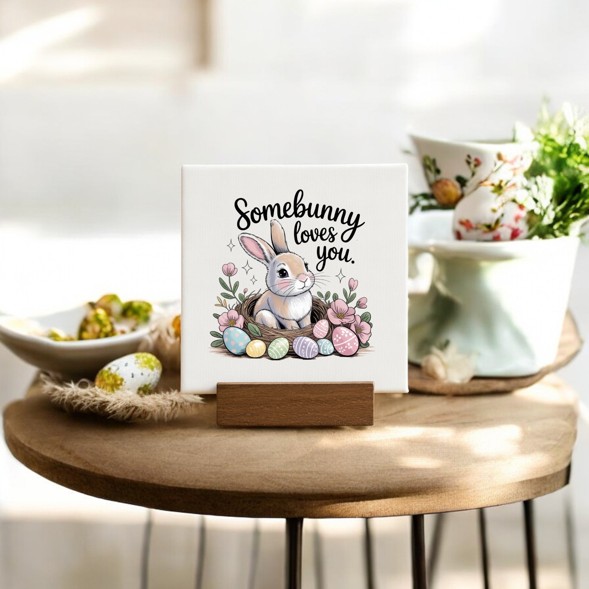 The Mirabilia Boutique "Somebunny Loves You" Mini Canvas (4x4) with wood stand features a bunny, Easter eggs, and flowers. Ideal as an Easter basket filler or spring decor. Perfect for tables or shelves.