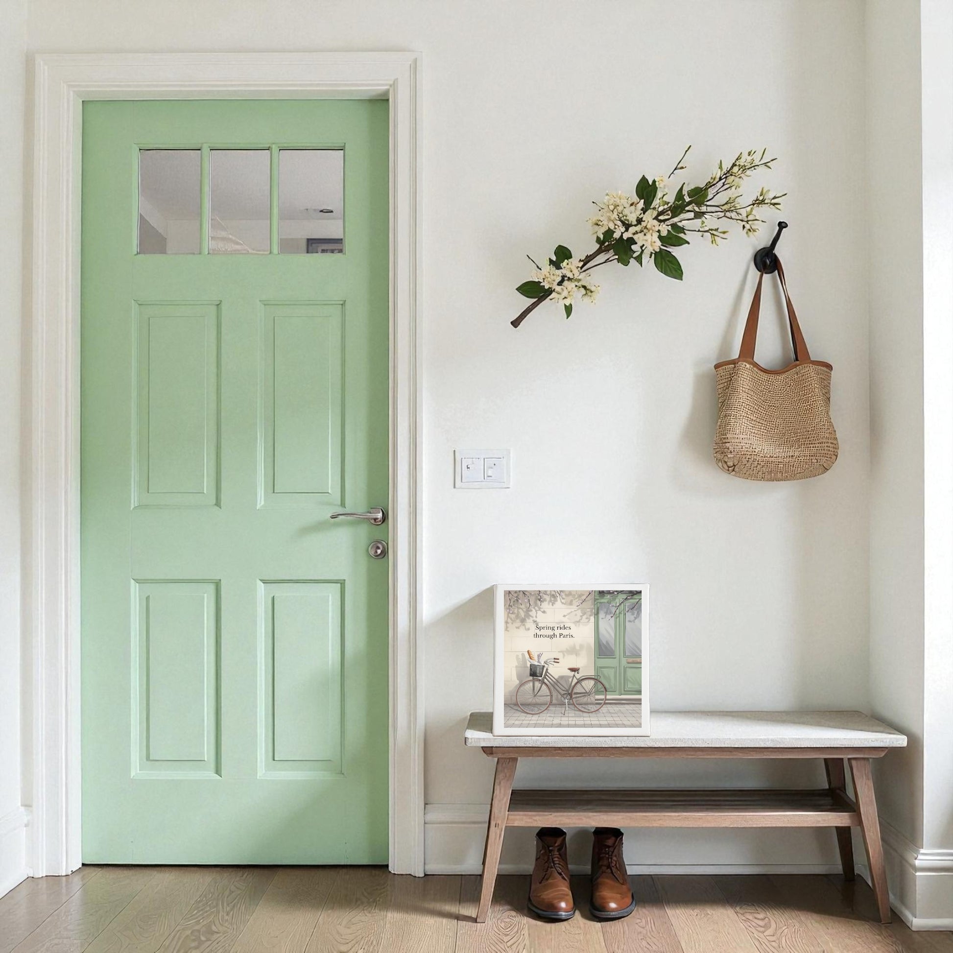 A light green door stands by a white wall with a hanging woven bag and flowering branch. Below, a bench displays framed art, including Mirabilia Boutique's Spring Rides Through Paris Canvas Art, and brown boots rest on the wooden floor.