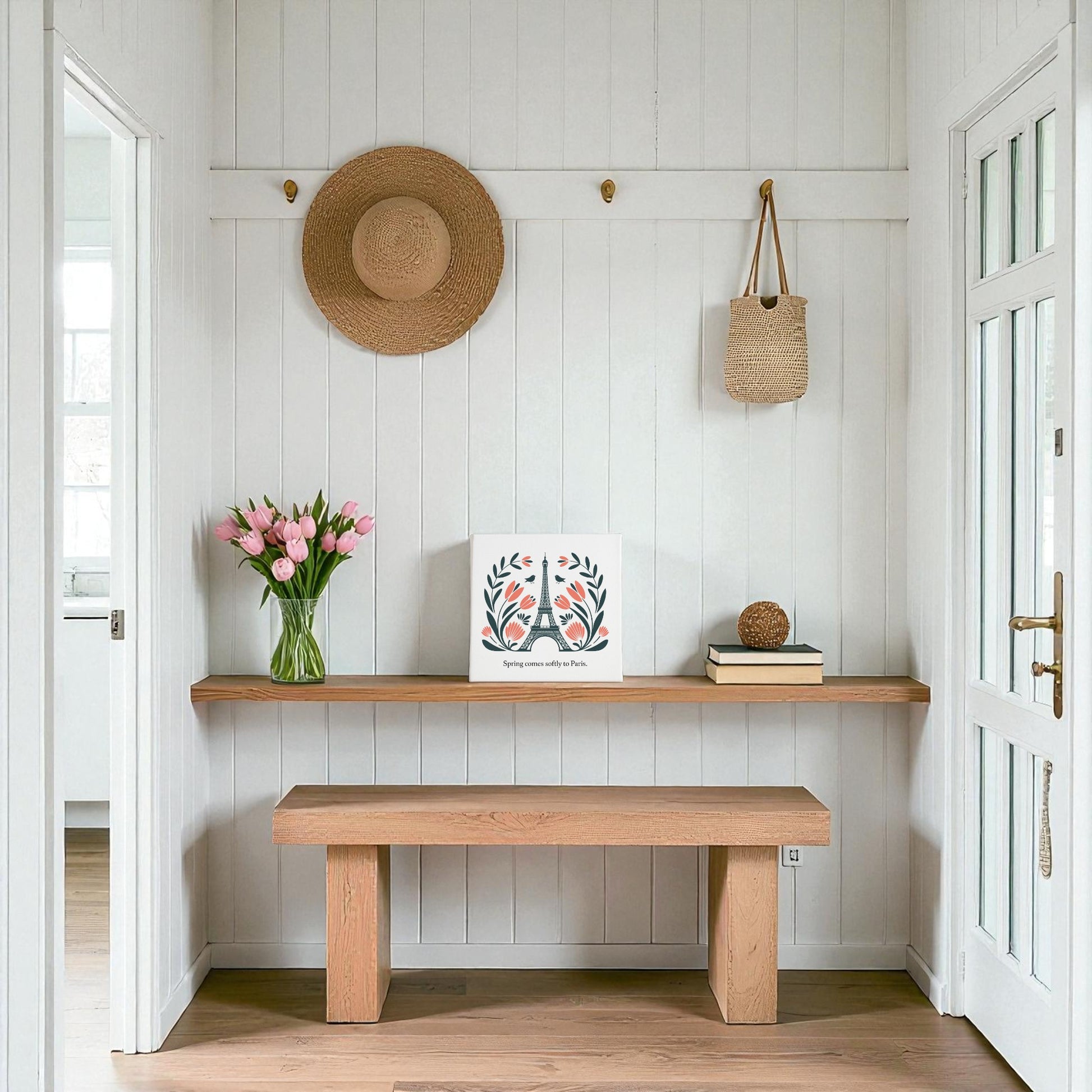 A bright entryway with white paneled walls, wooden bench, and shelf. On the shelf: pink tulips in a vase, Mirabilia Boutique's Spring Comes Softly to Paris Canvas Art, and a decorative ball. Hats and a straw bag hang above.