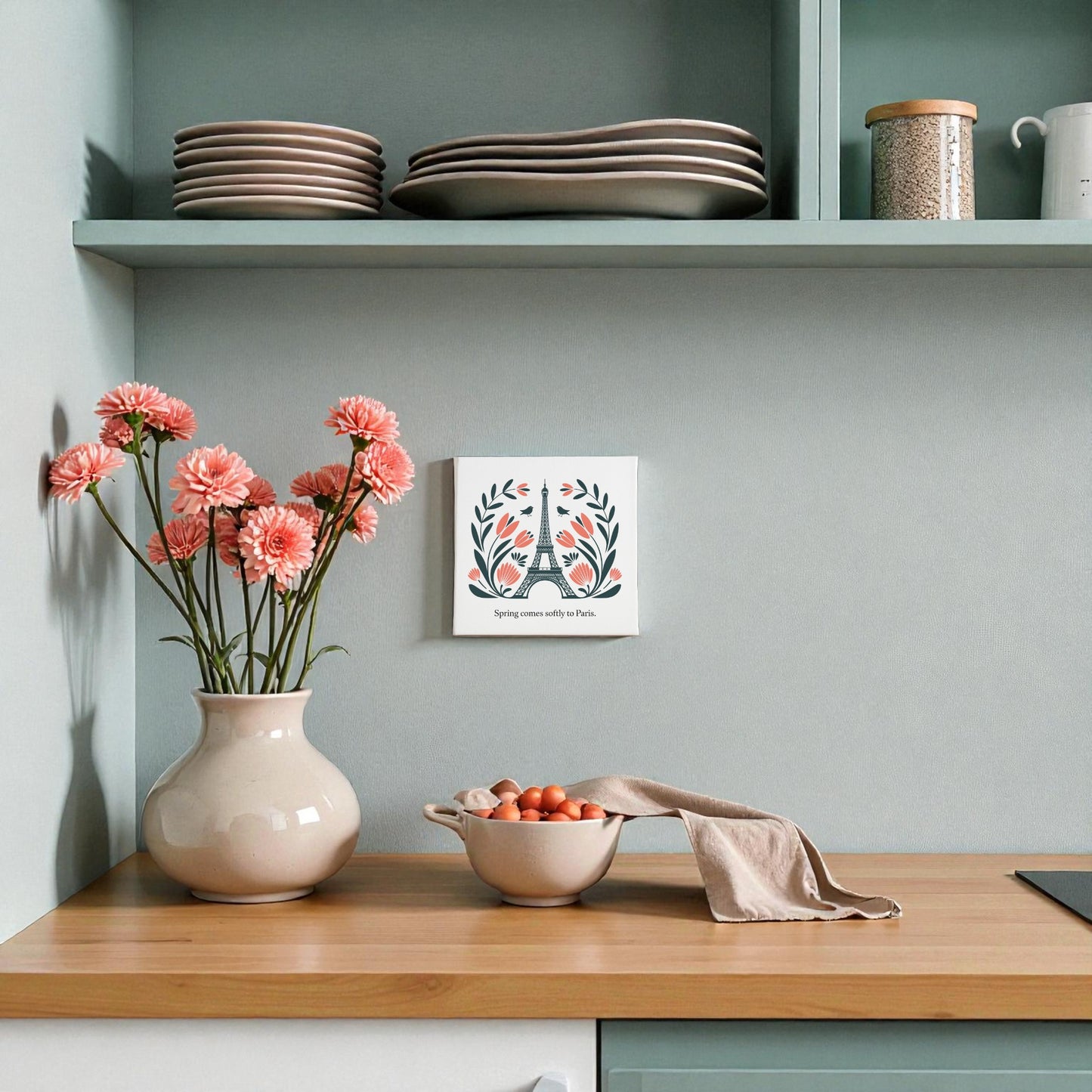 A kitchen counter features a vase of pink flowers, a bowl of tomatoes with a towel, stacked plates on a shelf, and Paris decor—Spring Comes Softly to Paris Canvas Art by Mirabilia Boutique adds charm with its floral folk art and motivational quote.