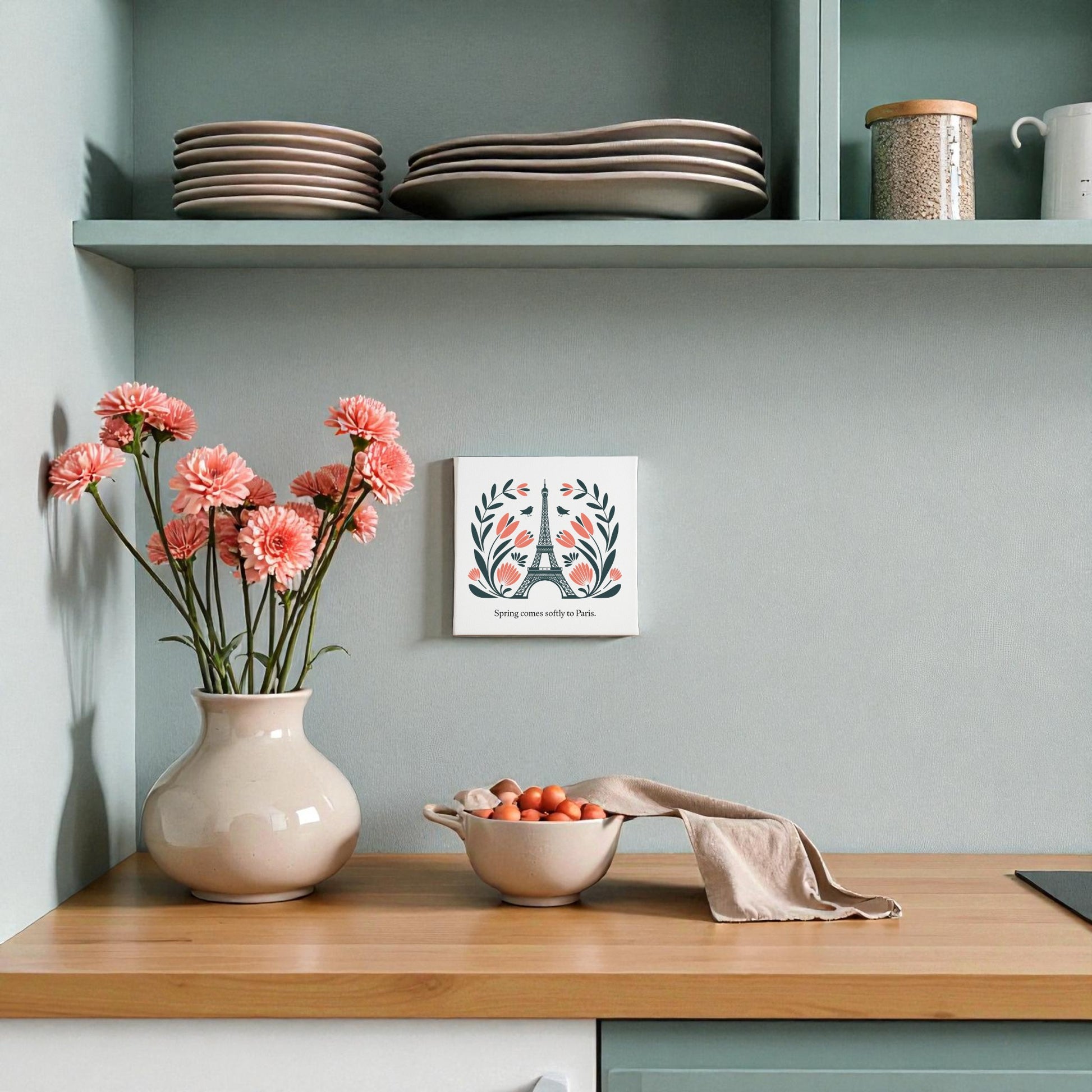 A kitchen counter features a vase of pink flowers, a bowl of tomatoes with a towel, stacked plates on a shelf, and Paris decor—Spring Comes Softly to Paris Canvas Art by Mirabilia Boutique adds charm with its floral folk art and motivational quote.