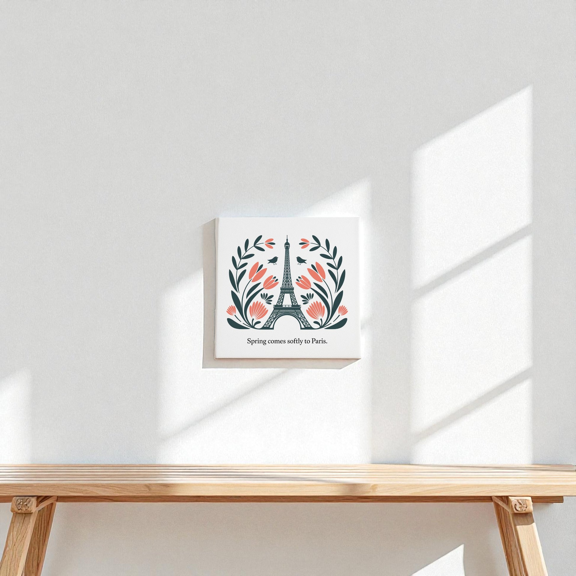 A small Spring Comes Softly to Paris Canvas Art by Mirabilia Boutique, featuring floral folk art of the Eiffel Tower, hangs on a white wall above a light wooden shelf, softly illuminated by sunlight and window shadows for elegant Parisian decor.