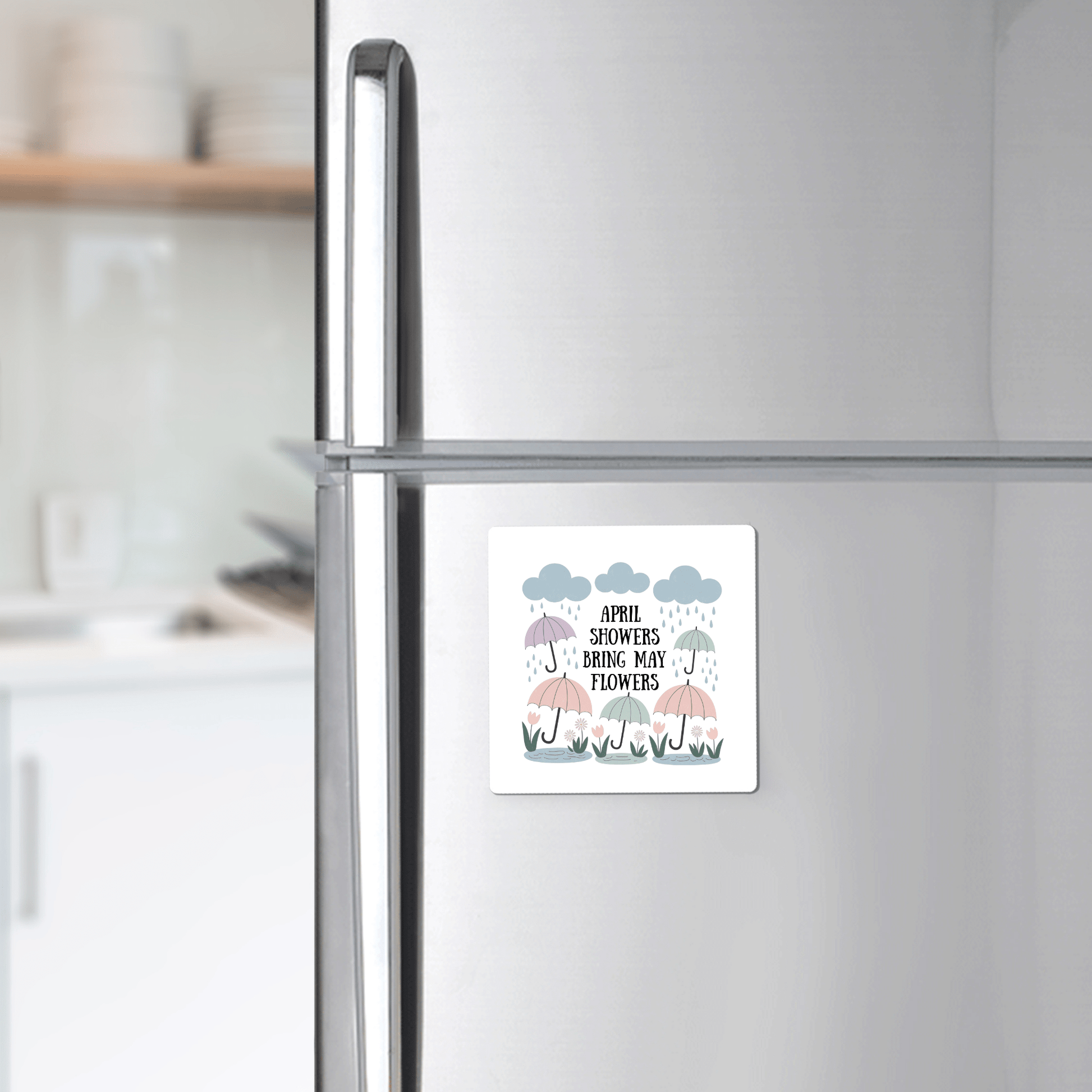 The April Showers Bring May Flowers Magnet by Mirabilia Boutique features pastel umbrellas, raindrops, and clouds on a square design, displayed on a silver fridge in a charming kitchen setting.
