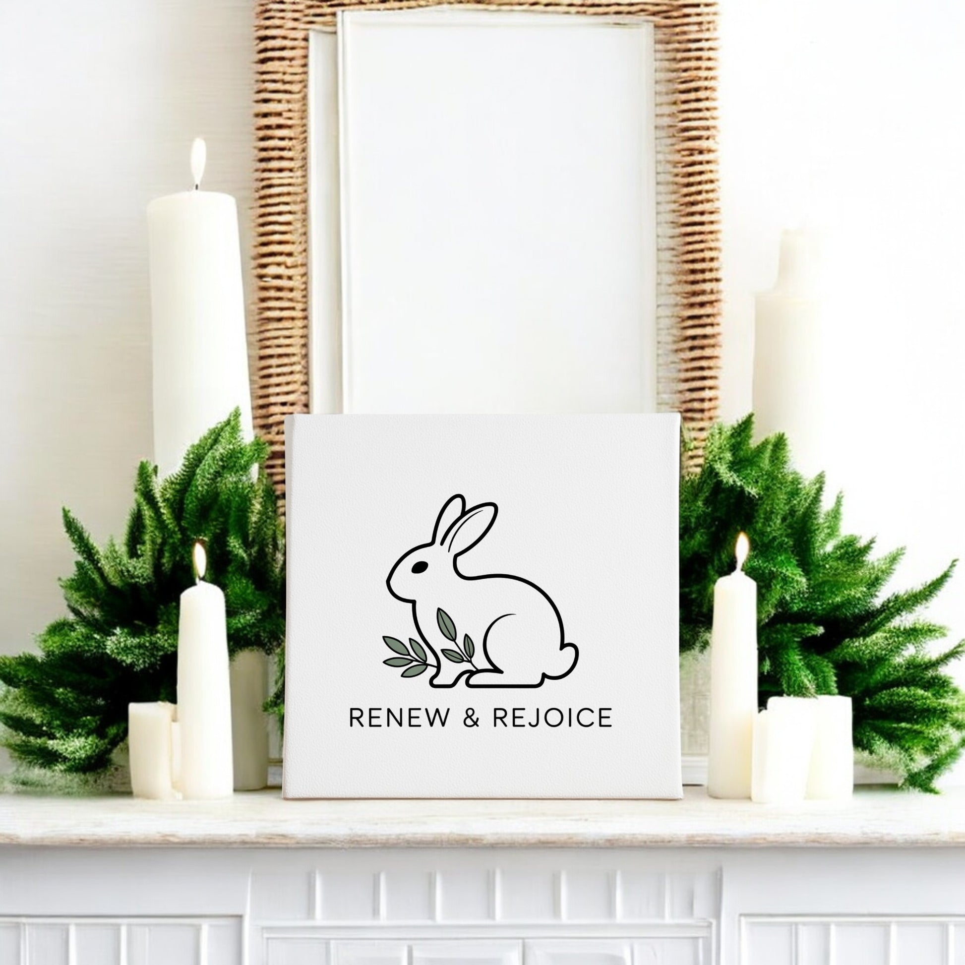A white mantel with green foliage and white candles displays the Mirabilia Boutique Renew & Rejoice Bunny Canvas – 8x8 Spring Wall Art, featuring “RENEW & REJOICE.” A woven mirror above completes this charming Easter decor.