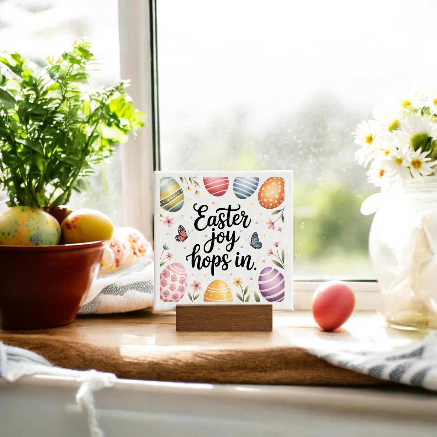 The Mirabilia Boutique Easter Joy Hops In Mini Canvas Sign (4x4) with colorful eggs and butterflies sits on a windowsill, surrounded by painted eggs, spring décor, a potted plant, and daisies soaking up the sunlight.